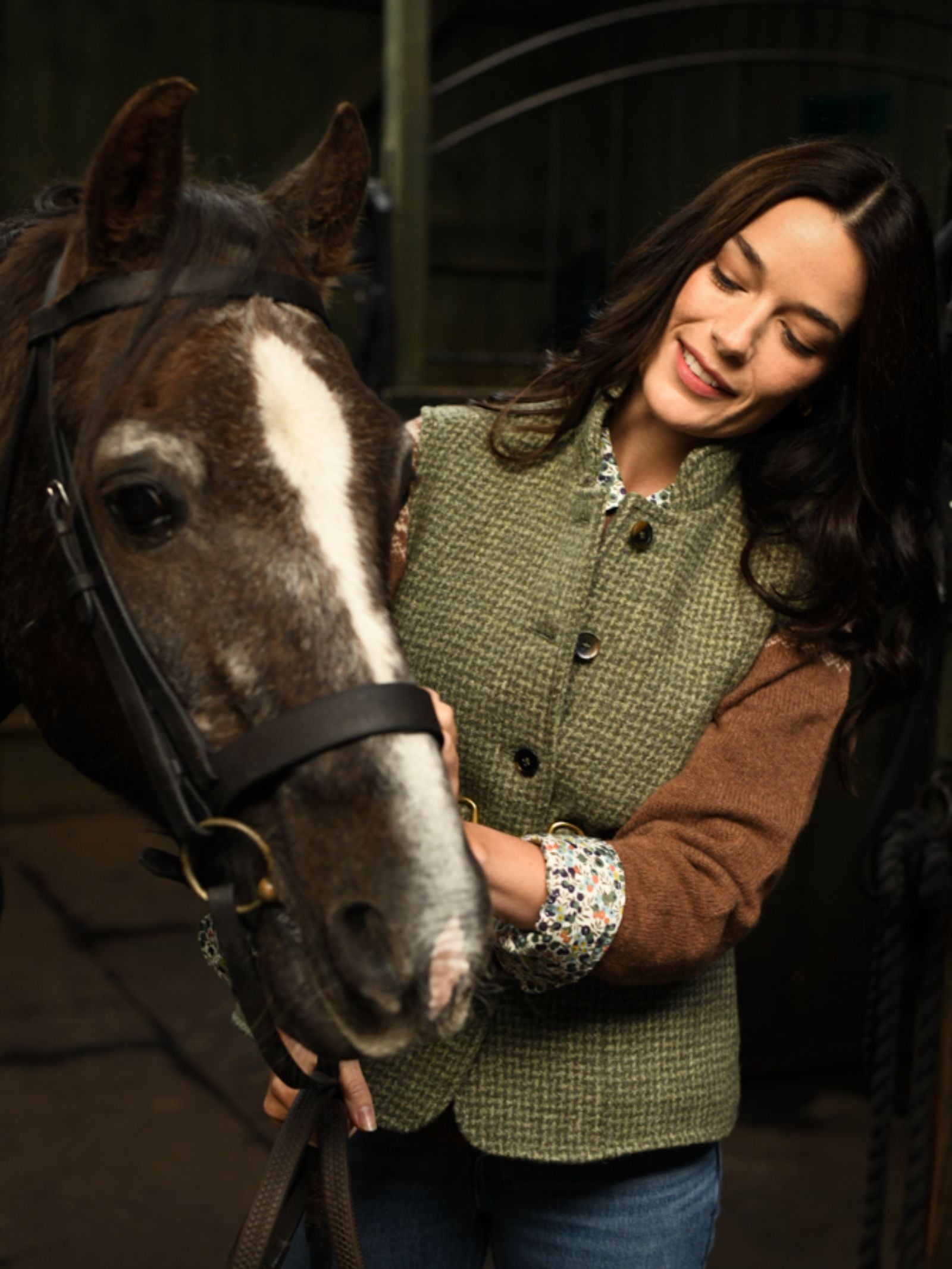 A woman with long dark hair, wearing the Campbell's of Beauly Auld Stock Harris Tweed Houndstooth Gilet, smiles as she gently pets a brown horse with a white stripe on its face in a stable.