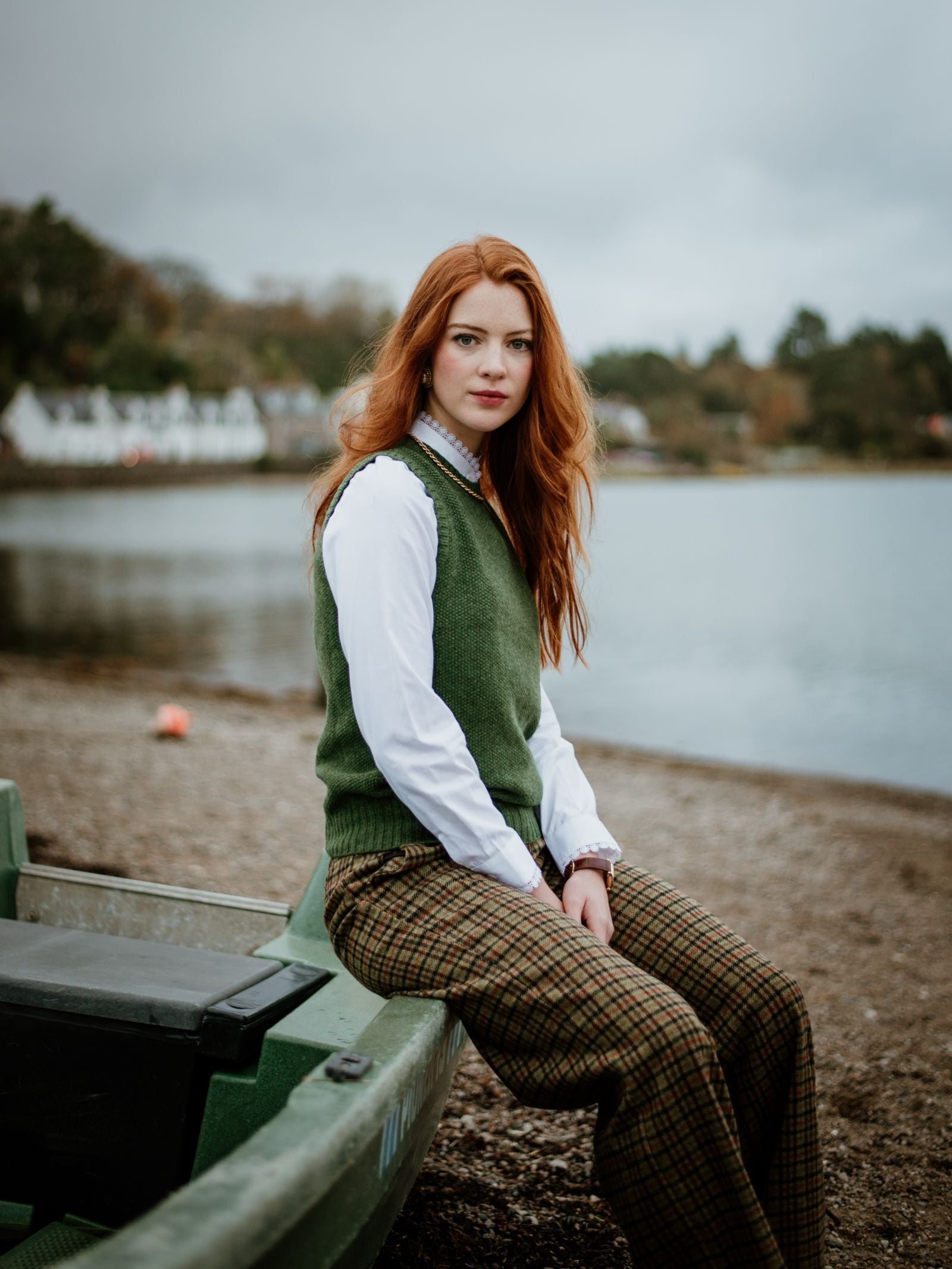 A young woman with long red hair, wearing the Campbells of Beauly Moss Stitch Crew Tank and plaid pants, sits on a green boat by a lake framed by trees and houses under a cloudy sky—a perfect setting for versatile layering.