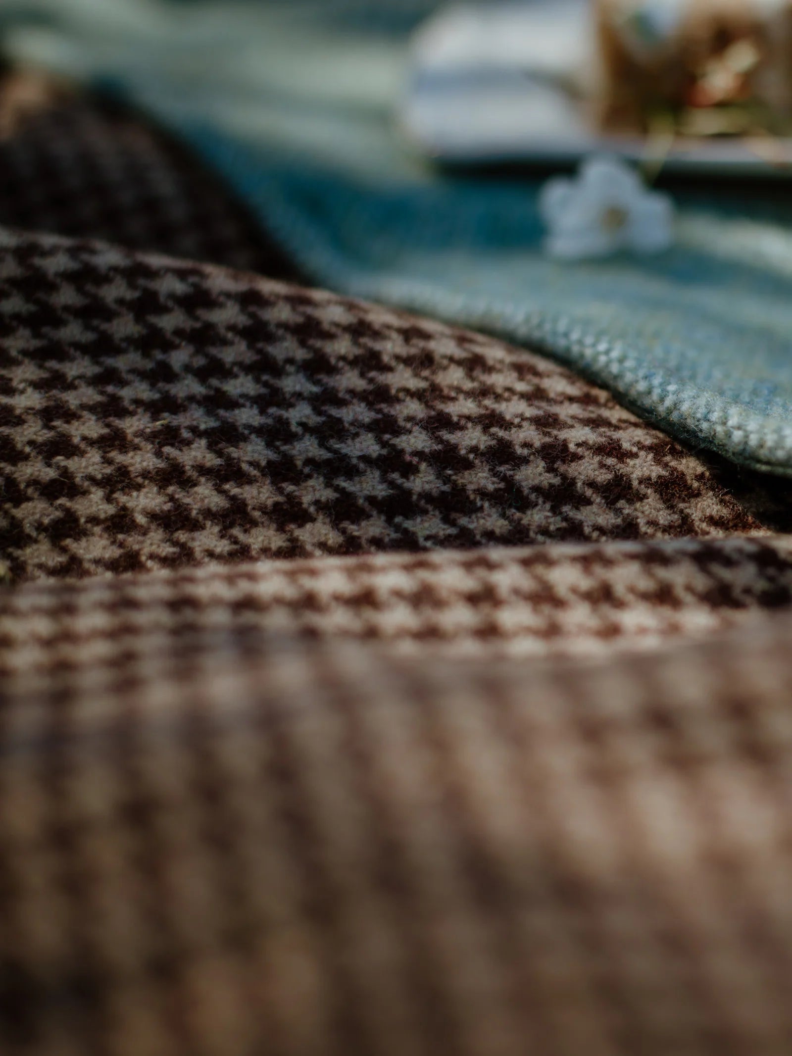 Close-up of the Crofters Blanket by Campbell's of Beauly, featuring a brown and beige houndstooth pattern. A green textured cloth and small white flower in the background add to its cozy, country charm.