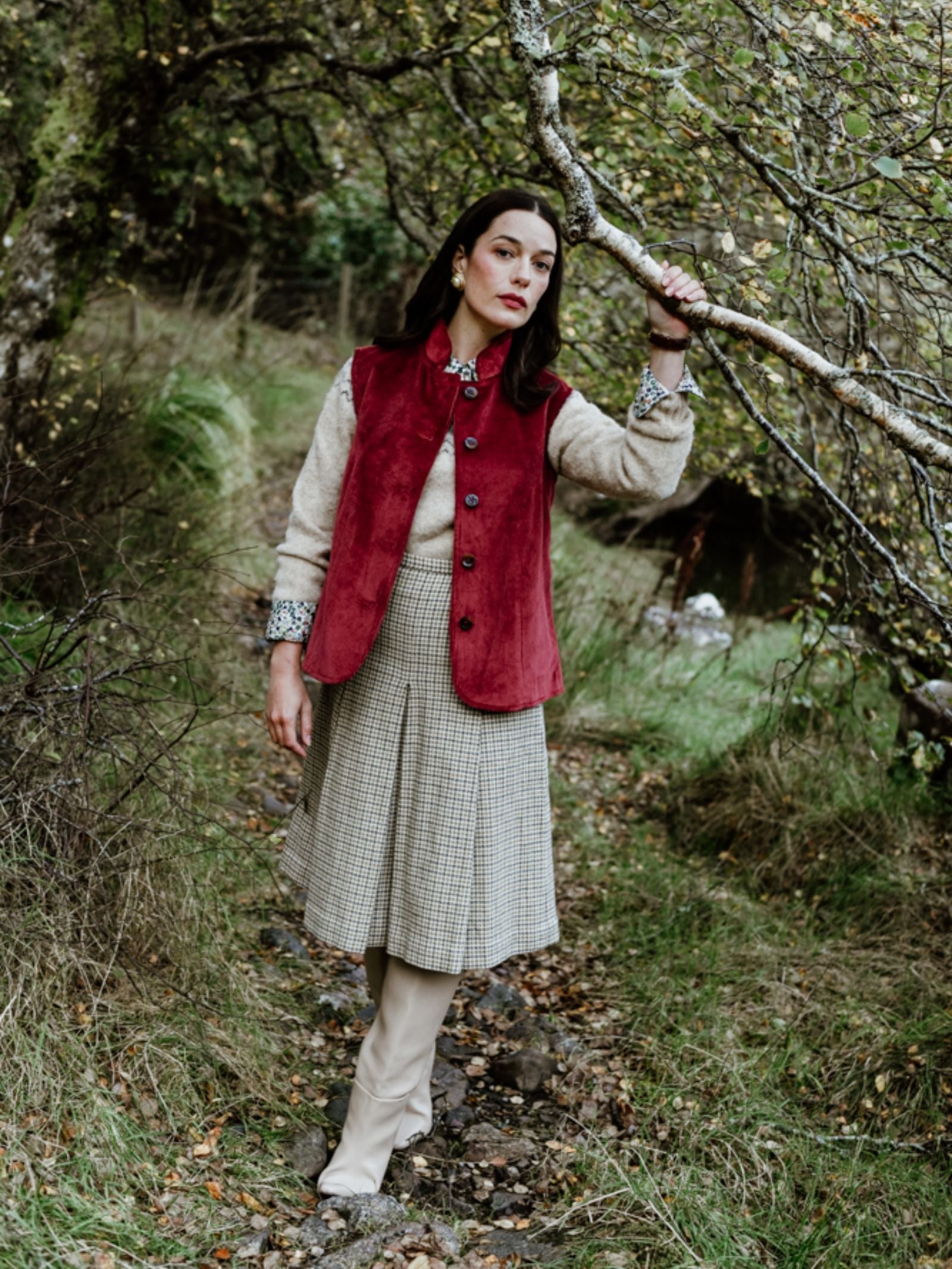 A woman stands on a forest path amid autumn leaves, wearing Campbell's of Beauly's Auld Stock Corduroy Gilet over a beige top, paired with a checked skirt and light boots, embodying timeless country elegance in nature.
