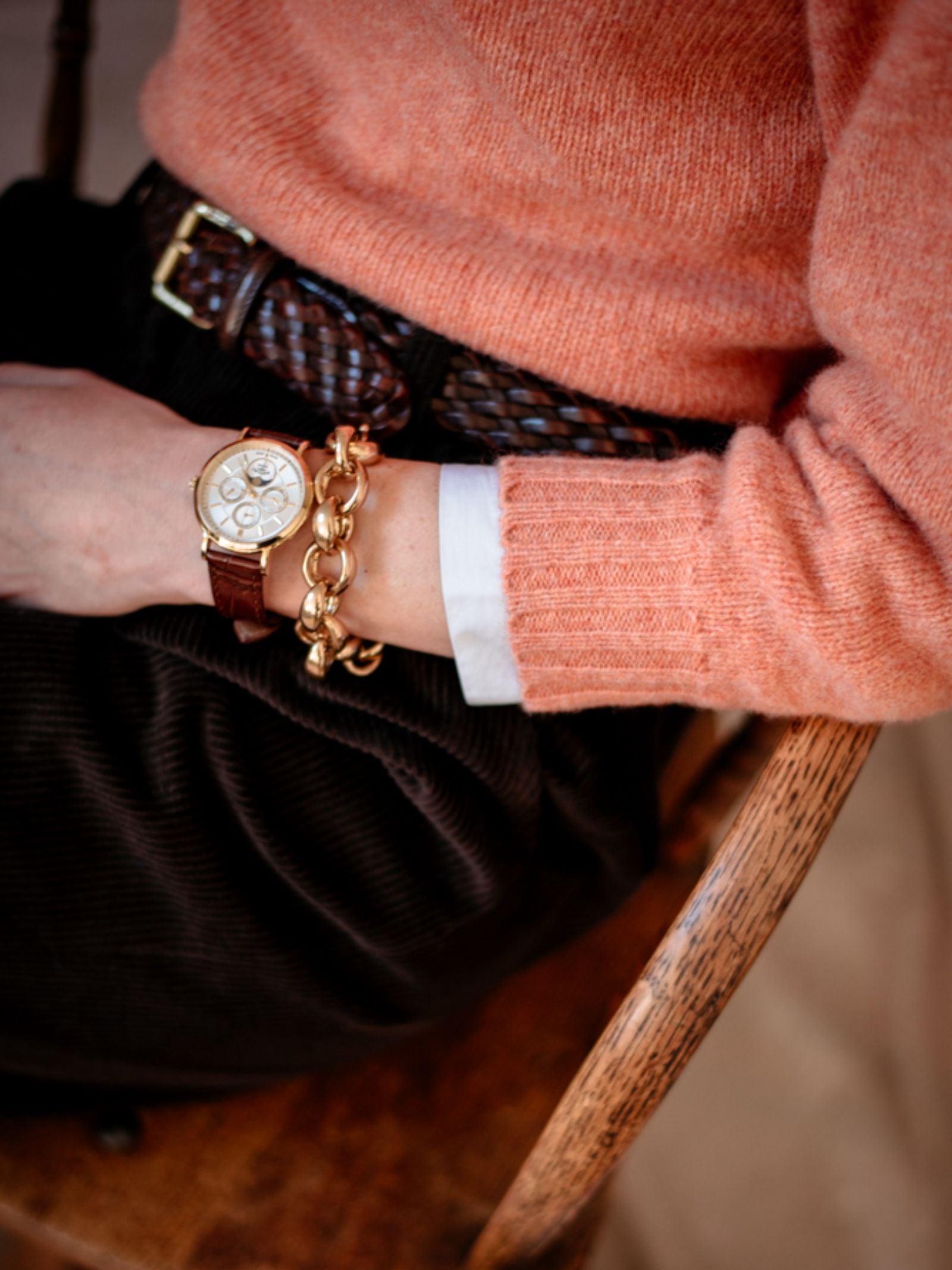 Seated on a wooden chair, a person wears the Campbell's of Beauly Auld Stock Cropped Shetland Jumper in peach over a white shirt, brown corduroy pants, and braided belt, accessorized with a gold watch and chunky gold chain bracelet.