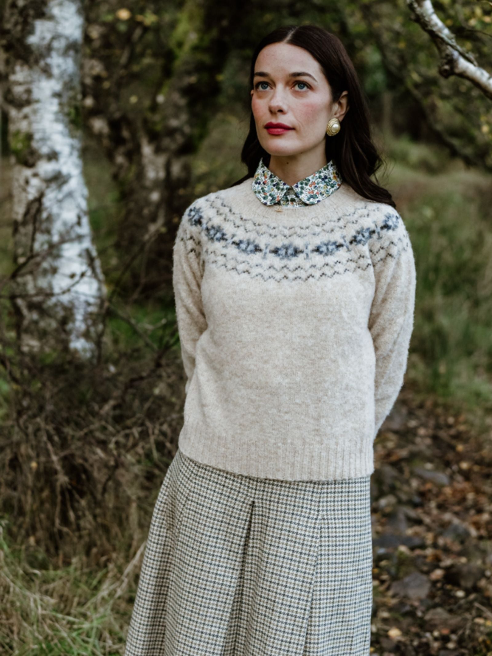 A woman with dark hair and gold earrings stands outdoors among trees, wearing Campbell’s of Beauly's Shaggy Cropped Fairisle Crew Jumper in Stone over a floral-collared shirt and checked skirt, looking serene in nature.