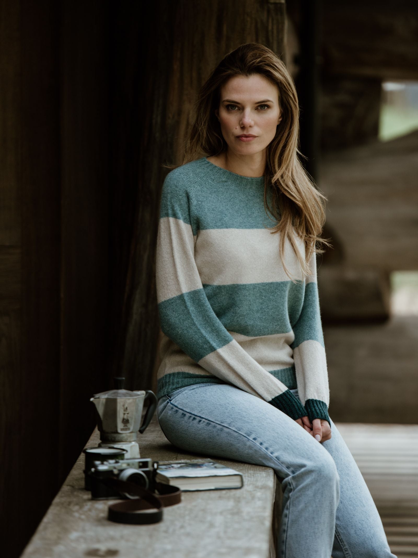 A woman with long hair wears the Campbell's of Beauly Auld Stock Stripe Crew Jumper and jeans, seated on a wooden ledge beside a coffee pot, camera, journal, and strap, looking calmly at the camera in a rustic setting.