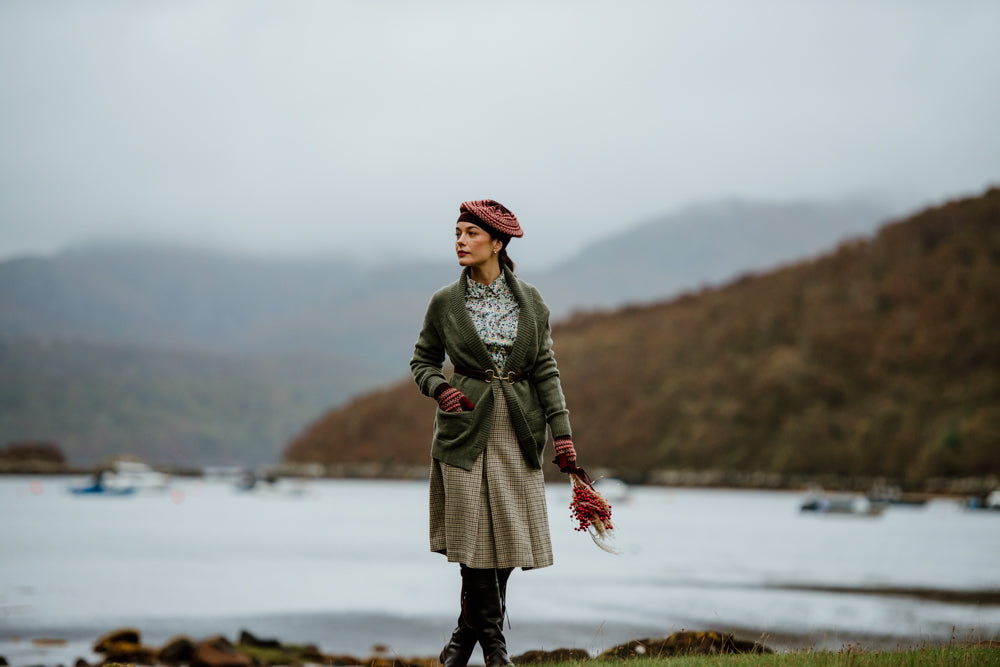 A woman stands by a misty lakeshore, wearing a green cardigan, patterned dress, maroon beret, and gloves. She holds a bouquet of red flowers, with hills and boats in the background.