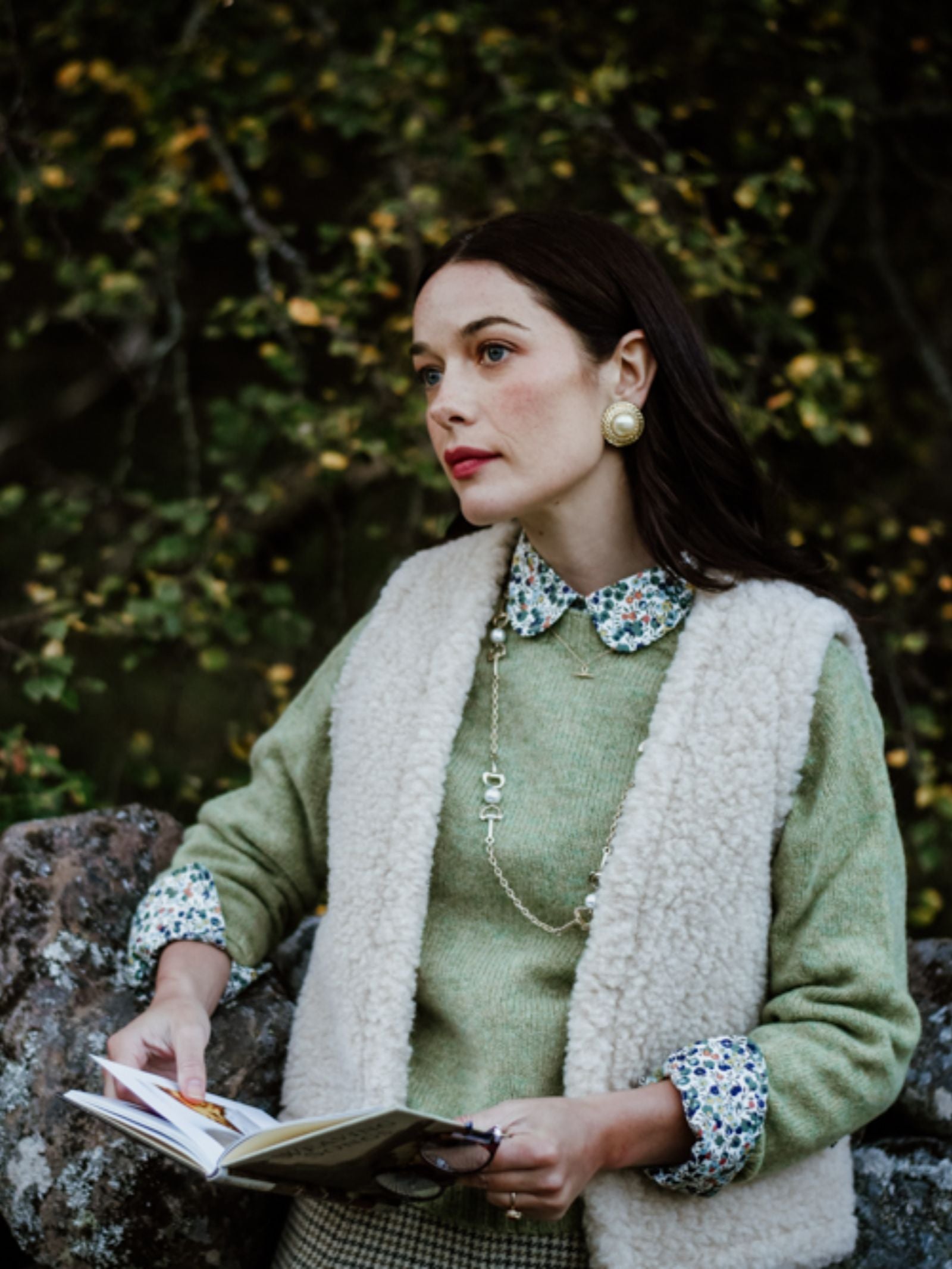 A woman with long brown hair stands outdoors by a stone wall, layering a Campbell's of Beauly Wool Shrug over a green sweater for lightweight warmth, paired with a plaid skirt, gold jewelry, and cream sherpa vest among the trees.