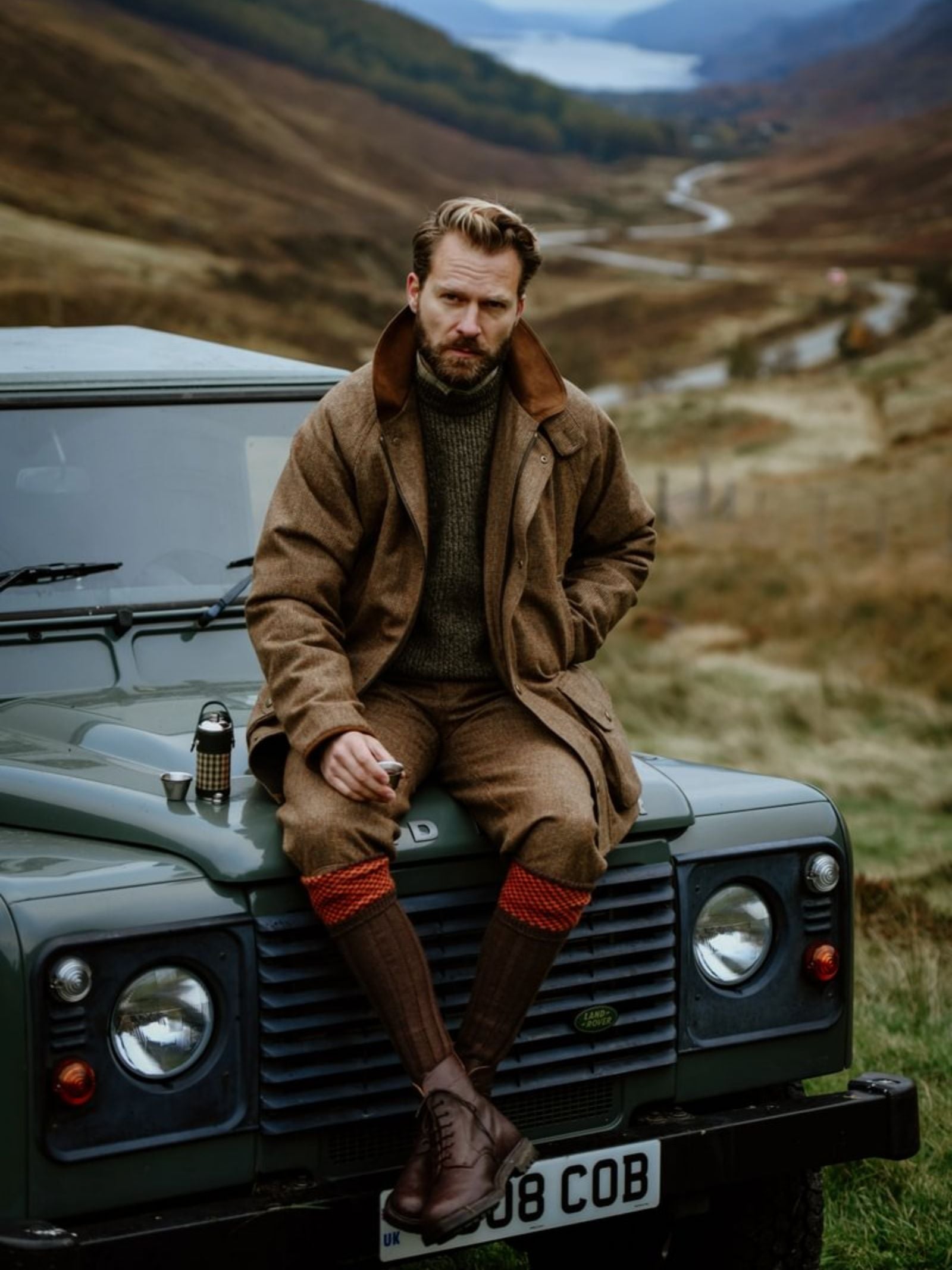 A man in brown outdoor clothing sits on a green Land Rover, holding a Campbell's of Beauly x Ettinger 8oz Forest Green Hunter Flask in a rural, hilly landscape with winding roads and a distant lake in the background.