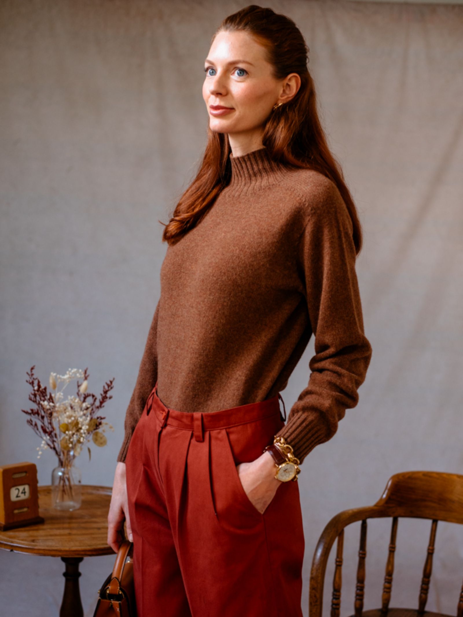 A woman with long reddish hair, wearing the Auld Stock Geelong Lambswool Funnel Neck Jumper by Campbell's of Beauly and rust-colored trousers, stands indoors with her hand in her pocket. A table with dried flowers and a wooden chair are behind her.