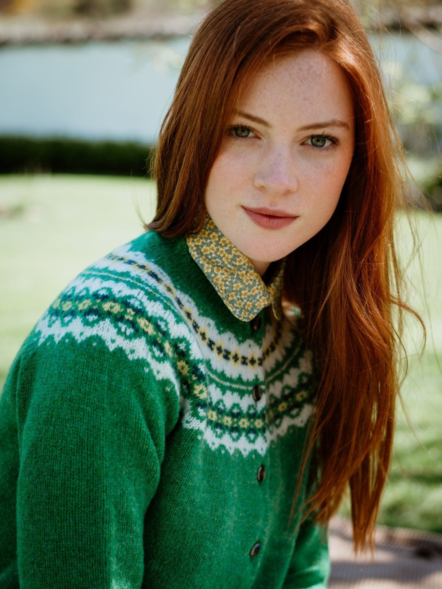 A young woman with long red hair and fair skin wears the Campbell's of Beauly Auld Stock Shetland Fairisle Cardigan over a floral collared shirt, sitting on grass and smiling slightly, embodying classic Scottish knitwear style.