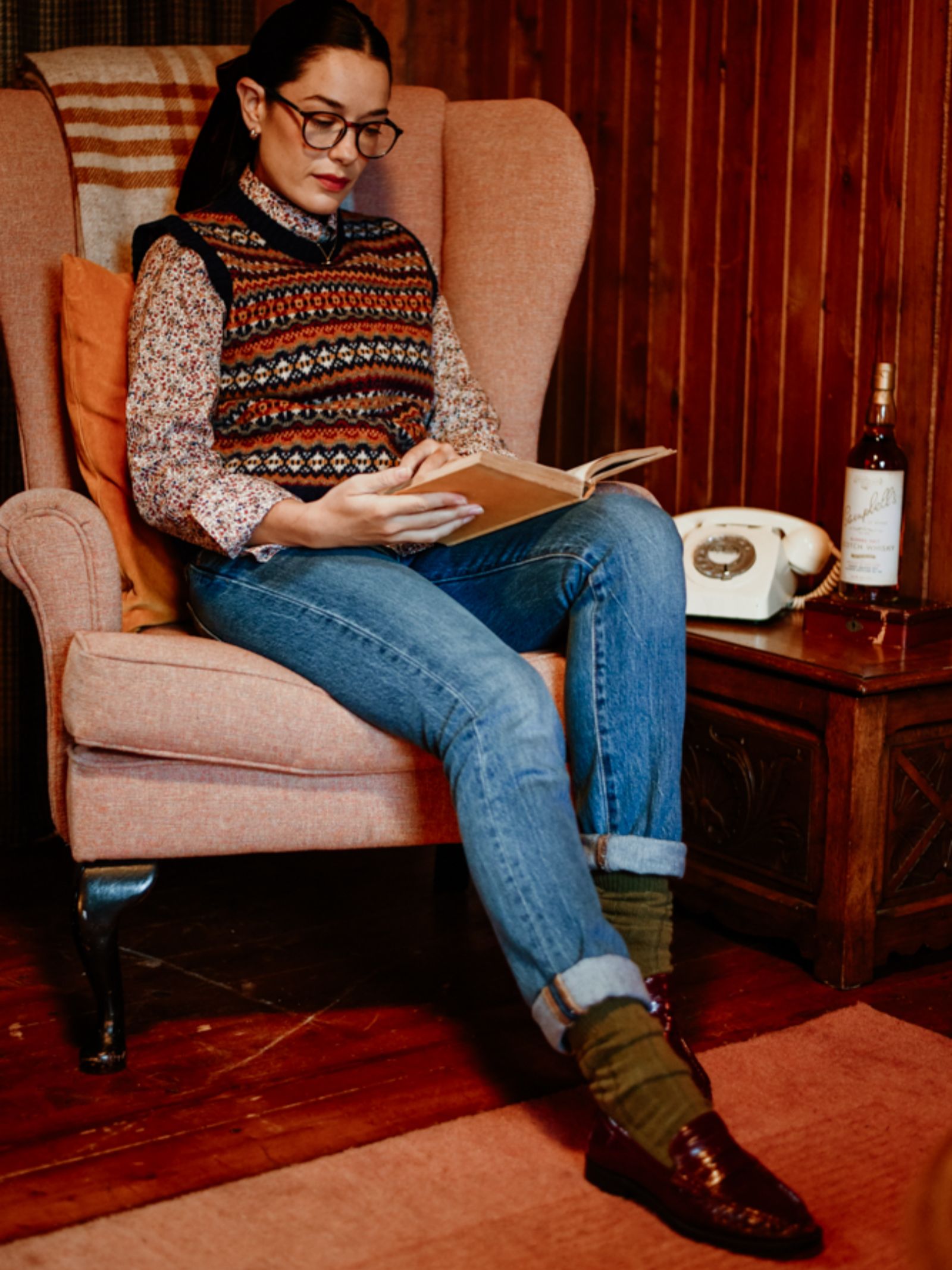 A woman wearing glasses, a patterned sweater vest, jeans, and Campbell's of Beauly Rib Cashmere Socks sits reading in an armchair. Next to her, a vintage phone and whisky rest on a wooden side table in a cozy, wood-paneled room.