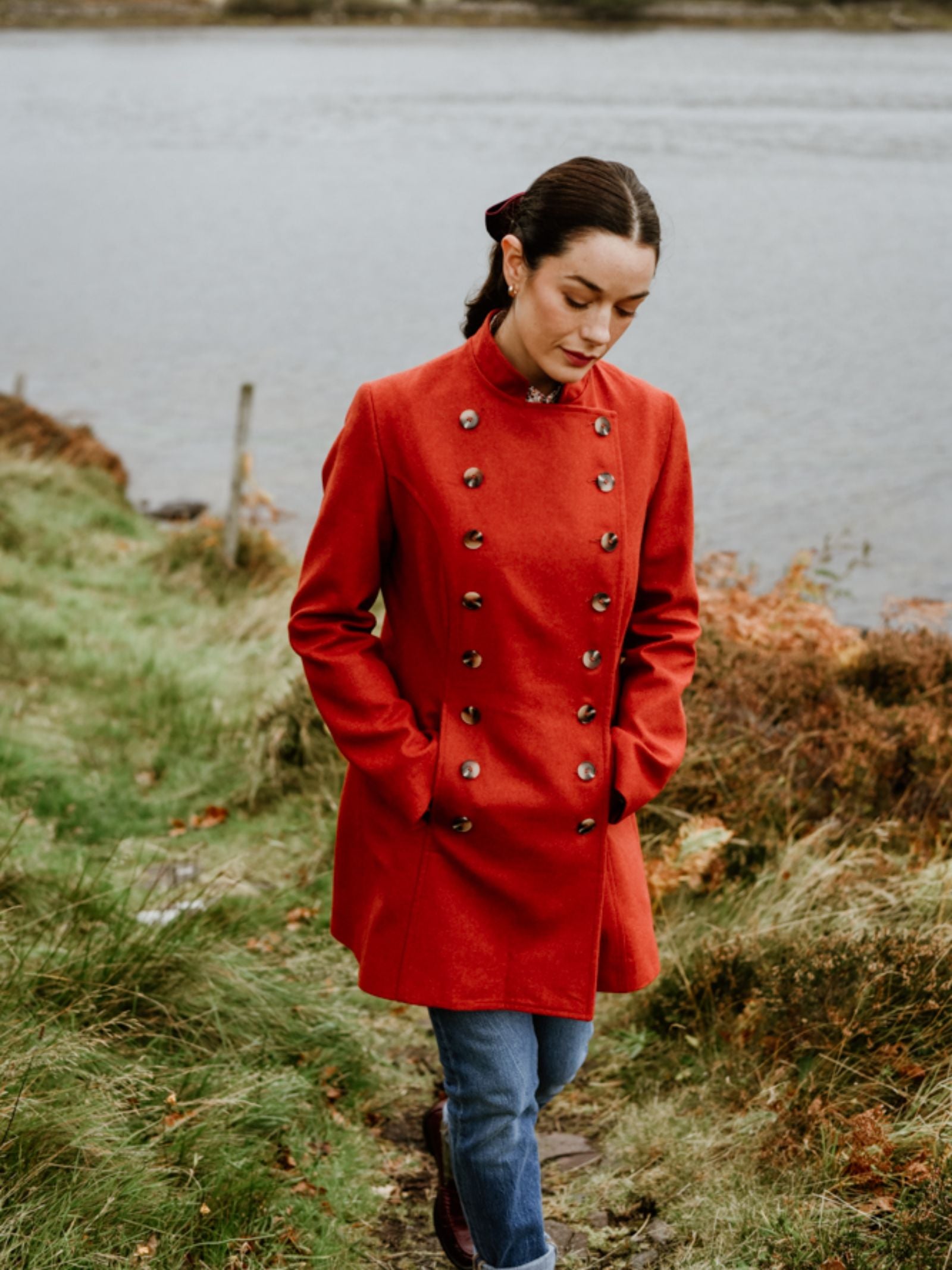 A woman wearing the Pirate Coat by Campbell's of Beauly strolls along a grassy path by the water, hands in her pockets and looking down with a serene expression.