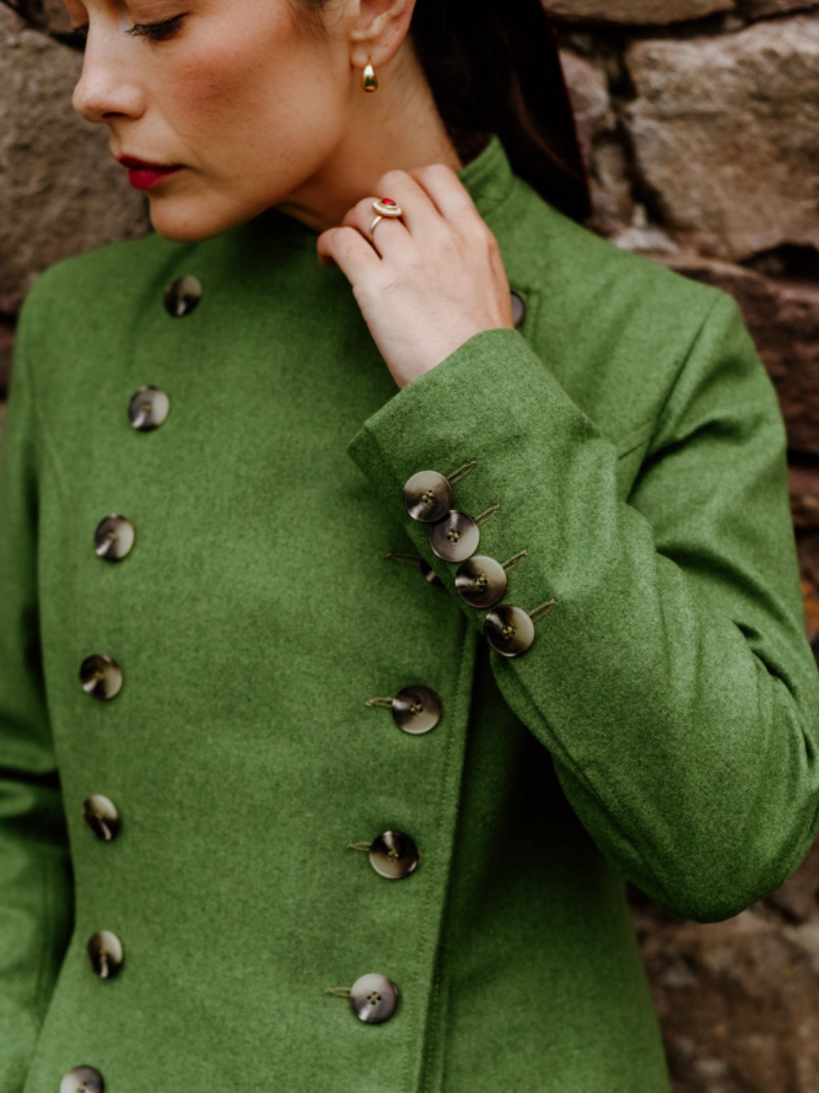 A woman in a green Campbell's of Beauly Pirate Coat with large decorative buttons stands by a stone wall, wearing red lipstick, gold earrings, and a gold ring with a red stone, gazing downward with her hand near her chin.