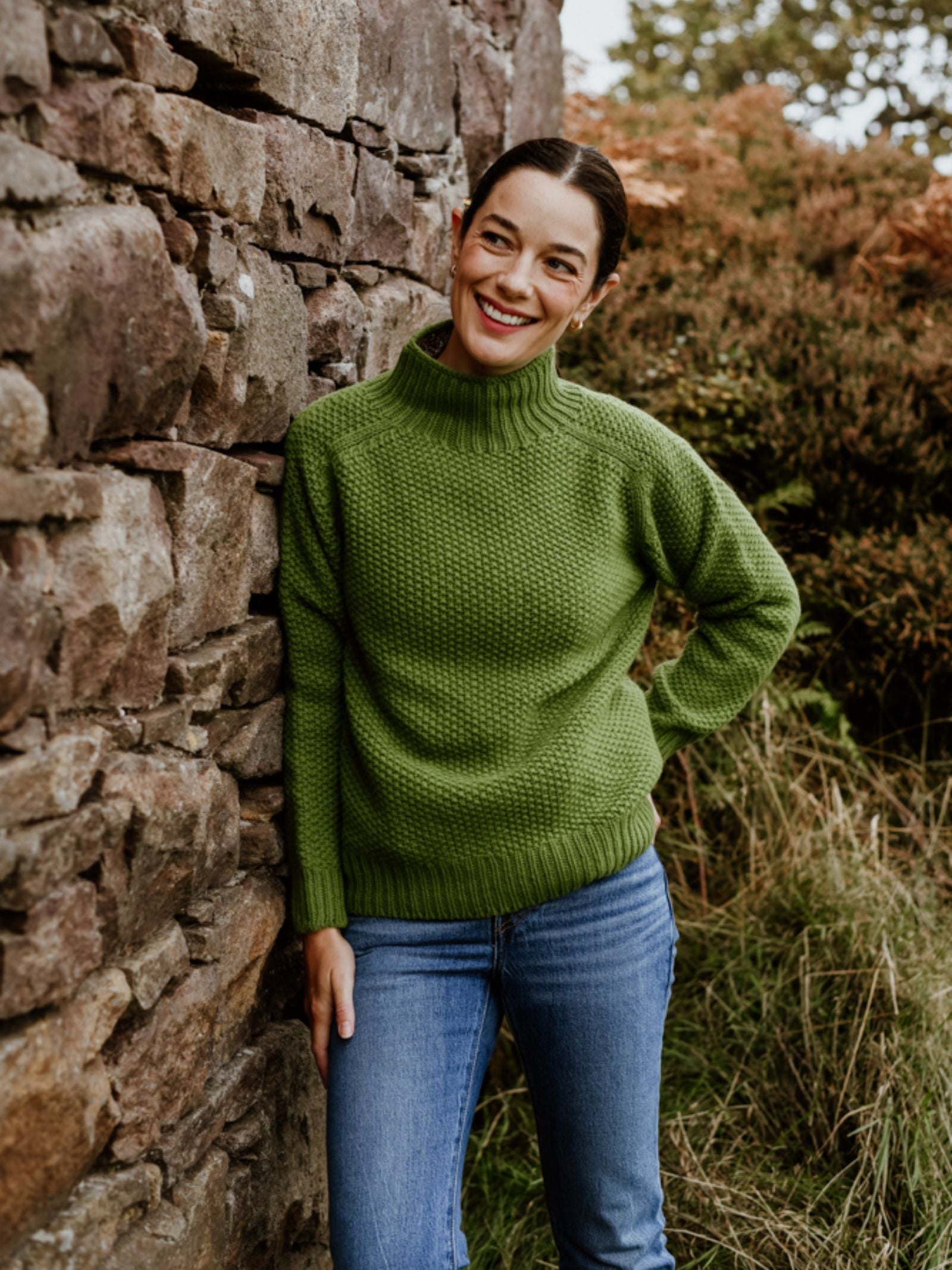 A person stands outdoors by a stone wall, smiling with one hand on their hip, wearing the Chunky Moss Stitch Polo by Campbell's of Beauly and blue jeans. Greenery and shrubs are visible in the background.