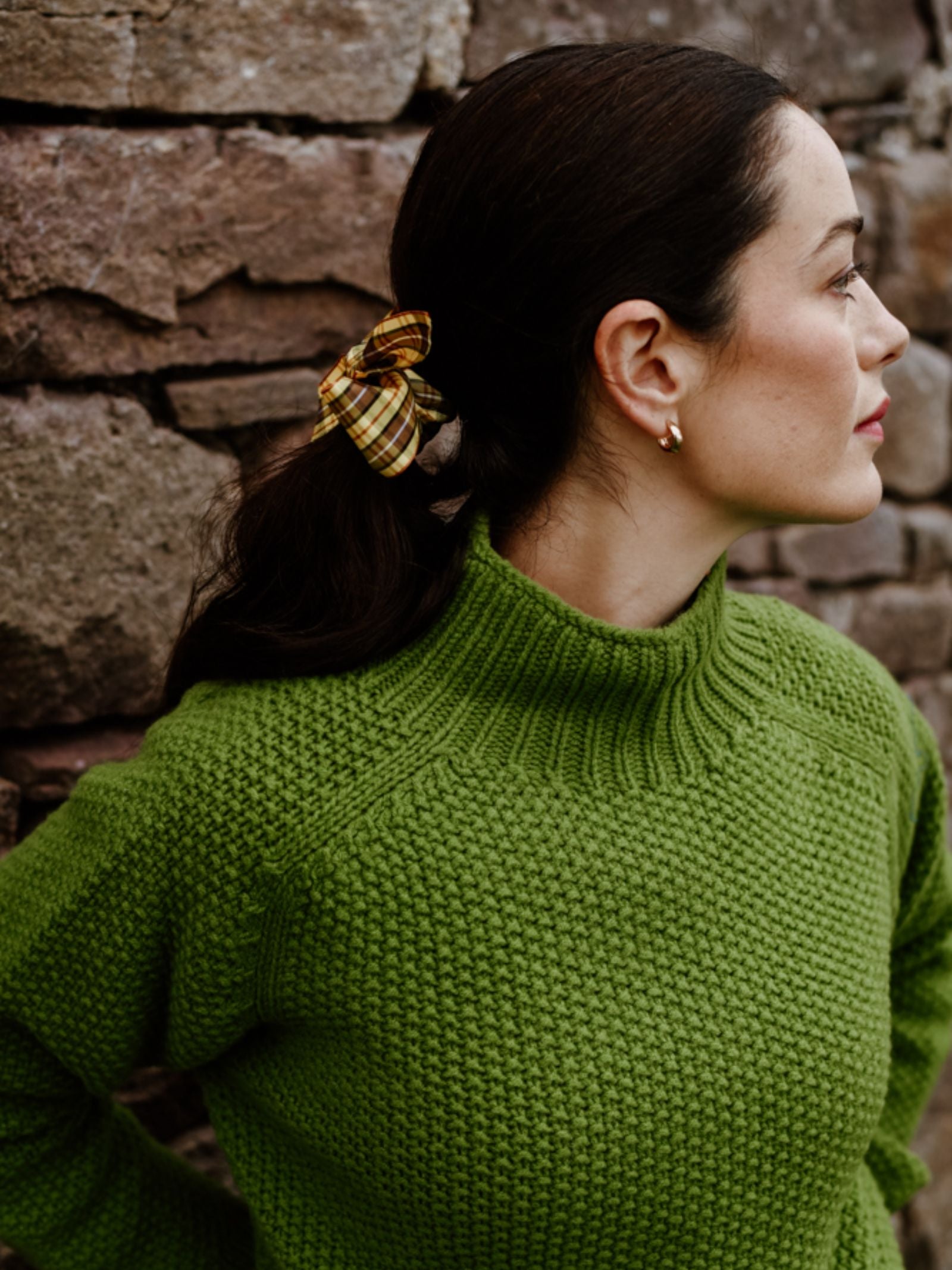 A woman with dark hair in a ponytail, tied with a yellow plaid scrunchie, wears Campbell's of Beauly's Chunky Moss Stitch Polo and gold hoop earrings, standing before a stone wall and gazing to the right.