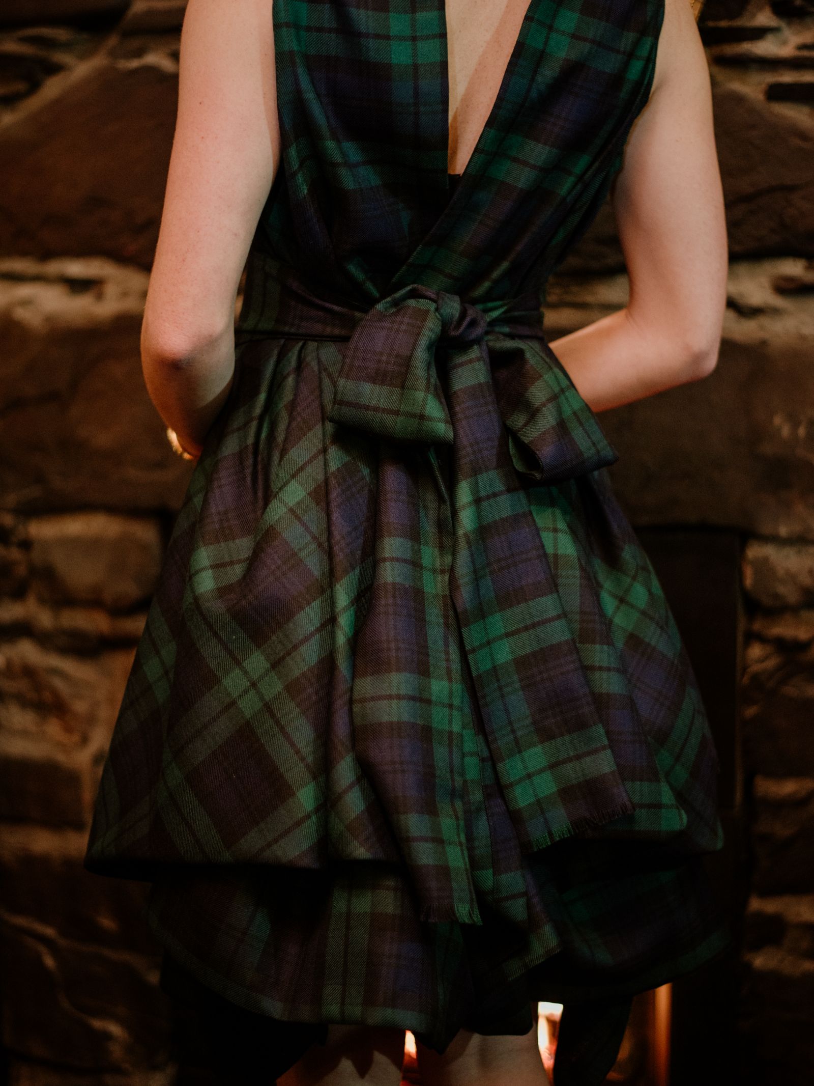 A person with light skin, seen from behind, wears a Campbell's of Beauly Reversible Wool Wrap Dress in green and navy tartan with a large bow at the back, standing before a stone wall.