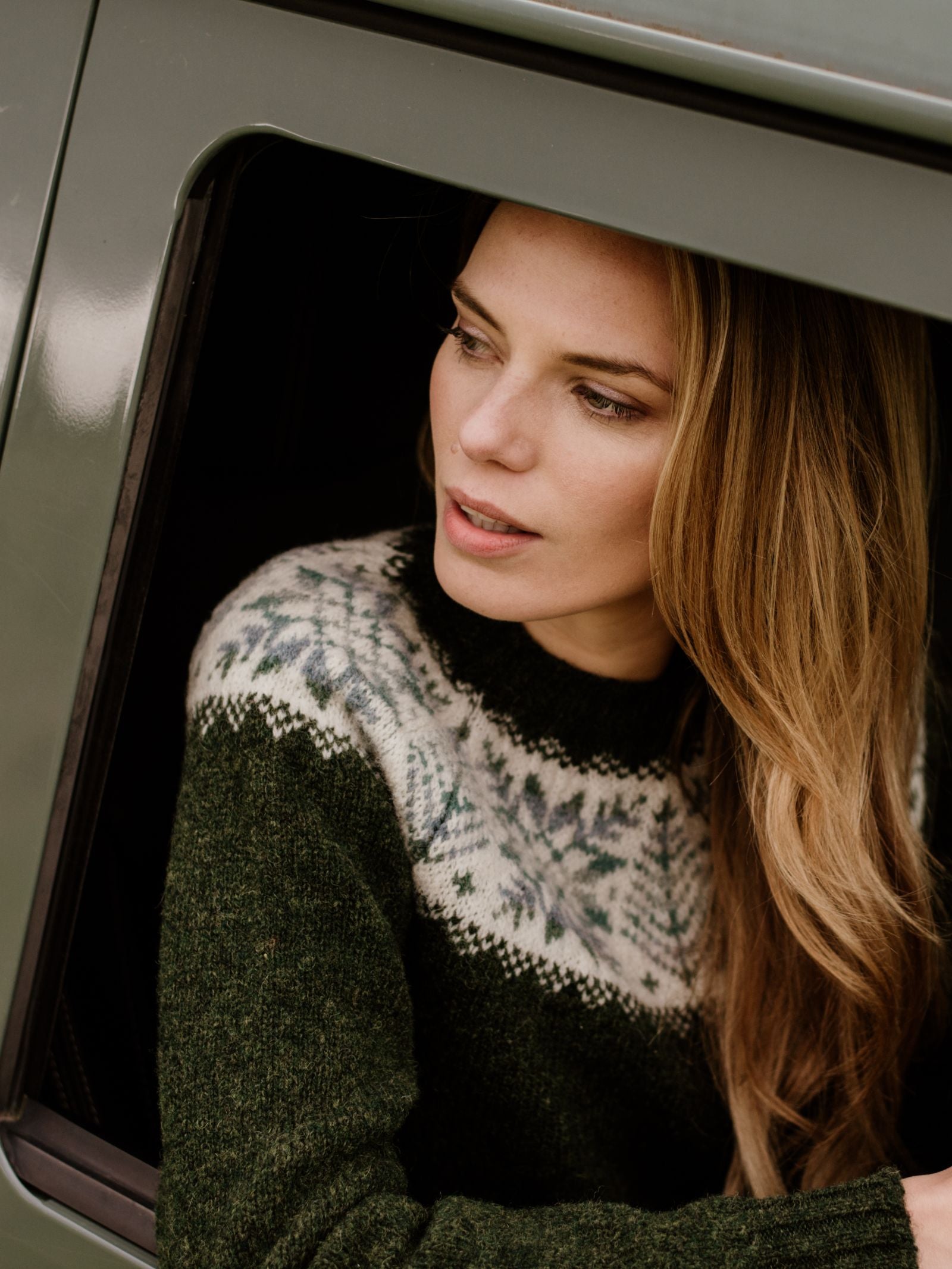 A woman with long blonde hair wears the Campbell's of Beauly Auld Stock Shetland Wool Snowflake Crew Jumper, featuring a white and gray Fairisle pattern, as she looks thoughtfully out of an open window in a green car.