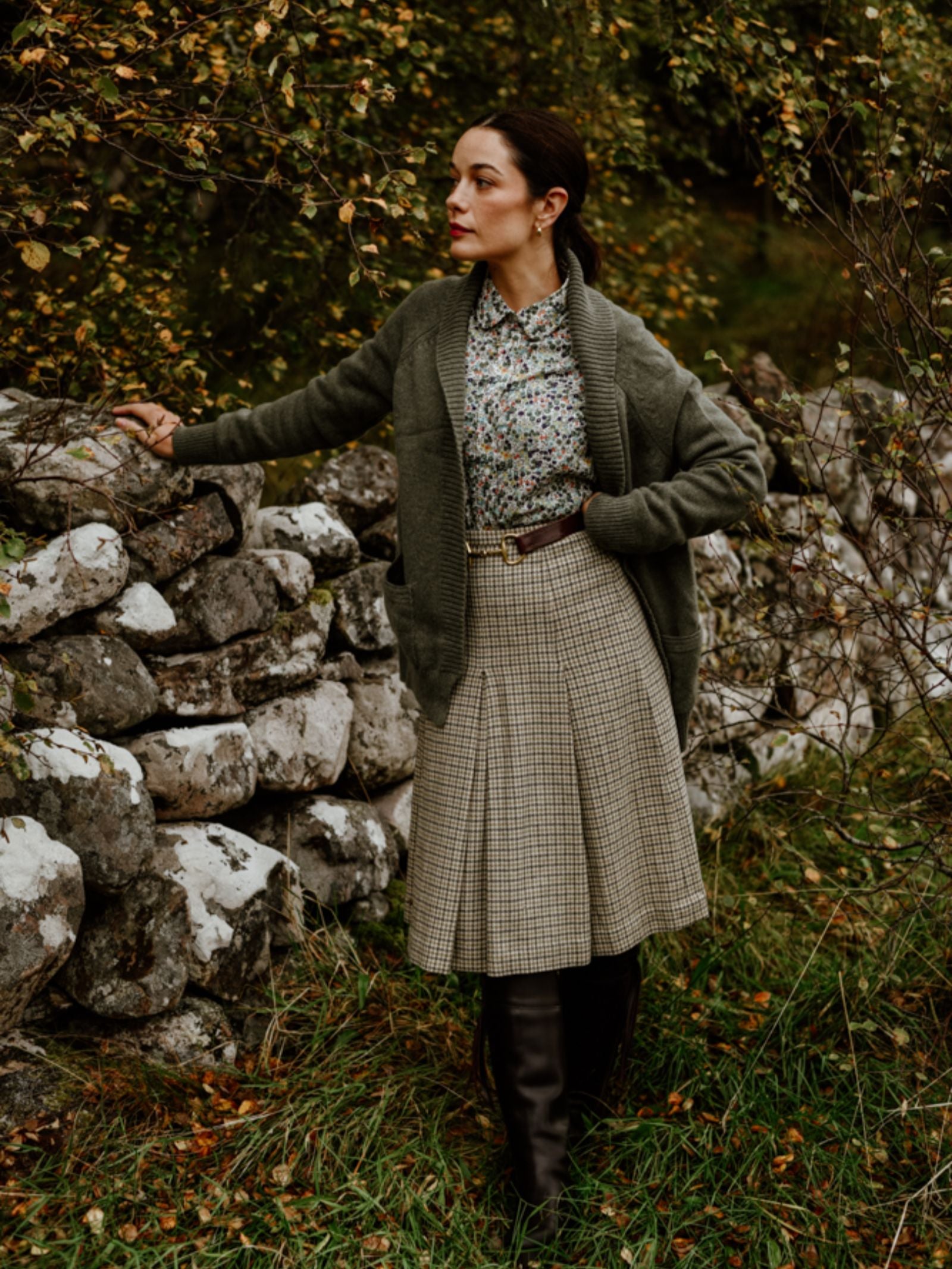 A woman stands by a stone wall, wearing a Campbell's of Beauly Cashmere Shawl Cardigan, floral blouse, plaid skirt, and tall black boots. Autumn leaves and grass surround her as she gazes thoughtfully into the distance.