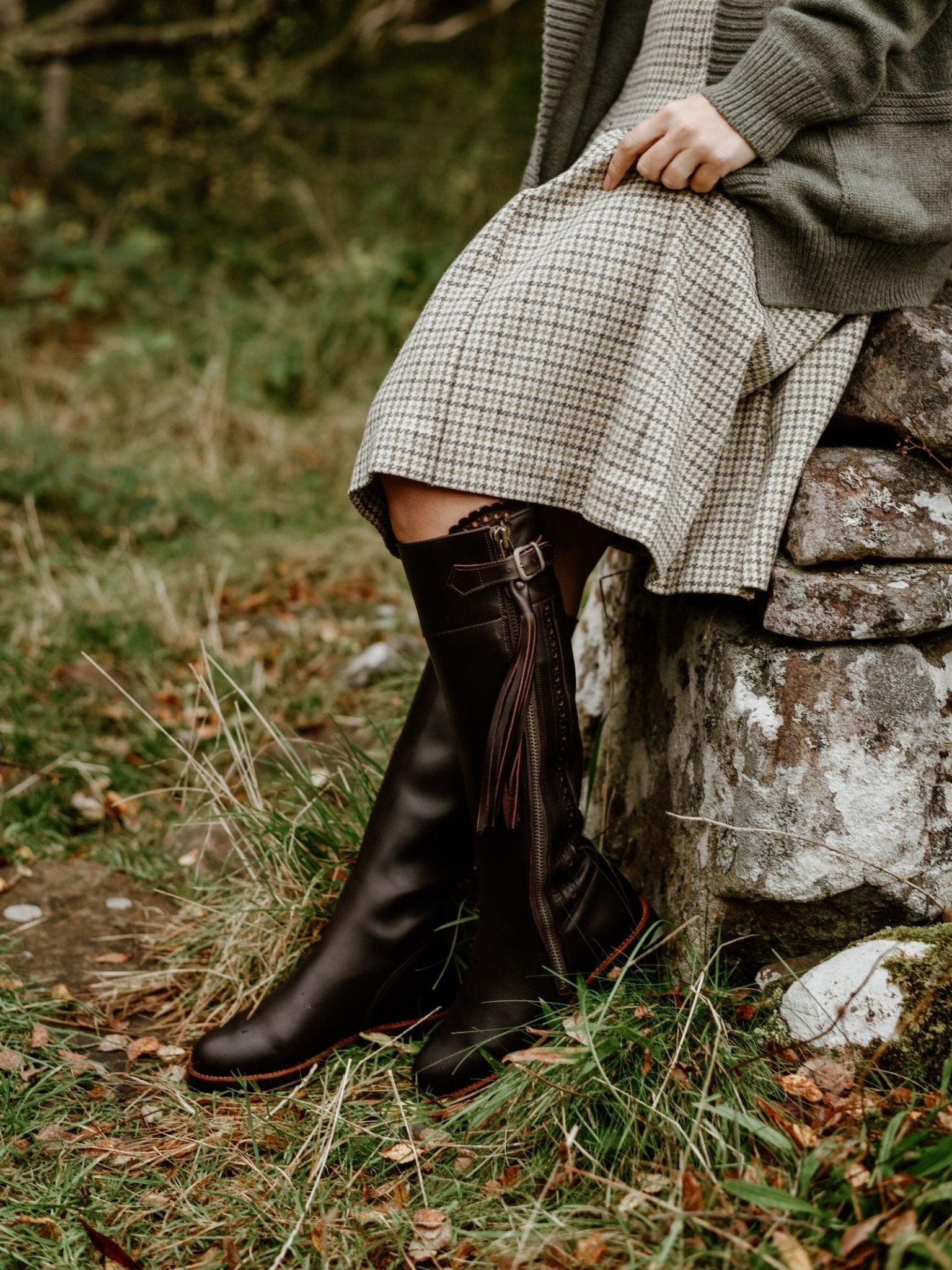 A person in a plaid dress, green cardigan, and tall dark brown Campbell's of Beauly Leather Spanish Boots with tassels sits on a stone wall in a grassy outdoor setting.