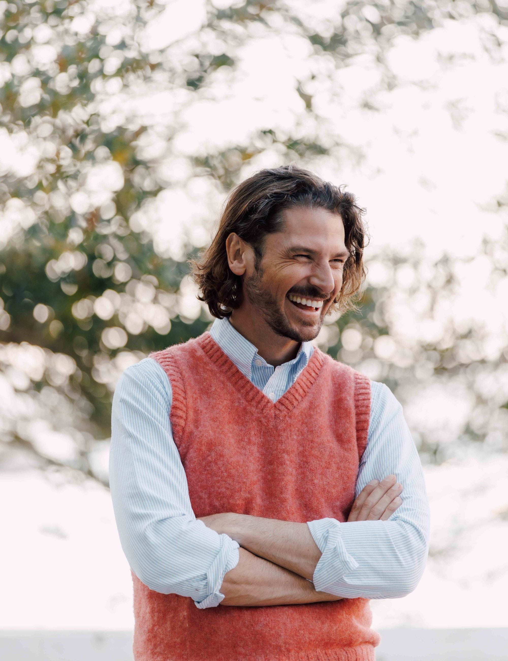A smiling man with wavy brown hair, wearing a pink knit vest over a light blue shirt, stands outdoors with his arms crossed in front of a blurred background of trees.