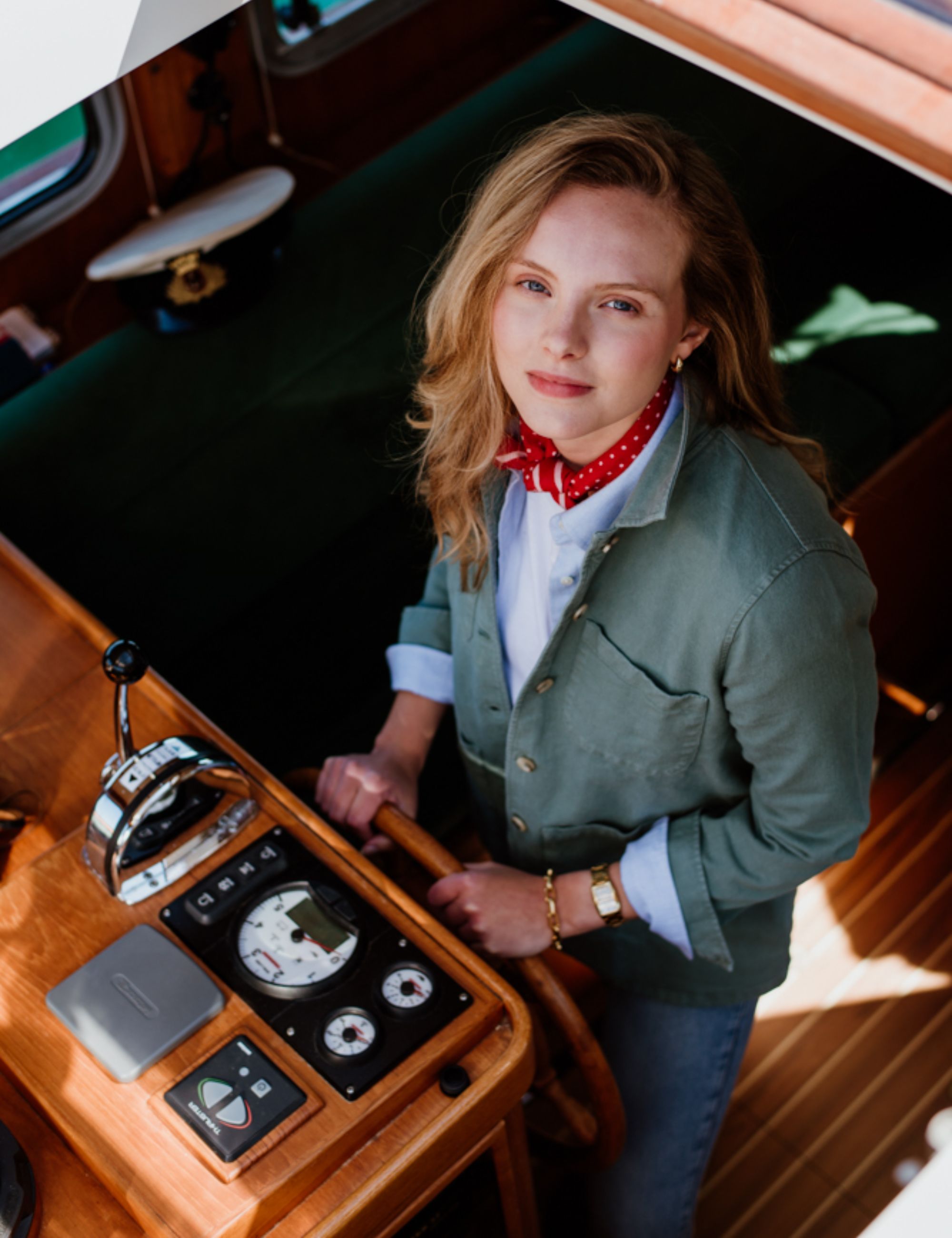 A woman with blonde hair, wearing a green jacket, red polka-dot scarf, and white shirt, stands at the helm of a wooden boat, looking up toward the camera with one hand on the steering wheel.