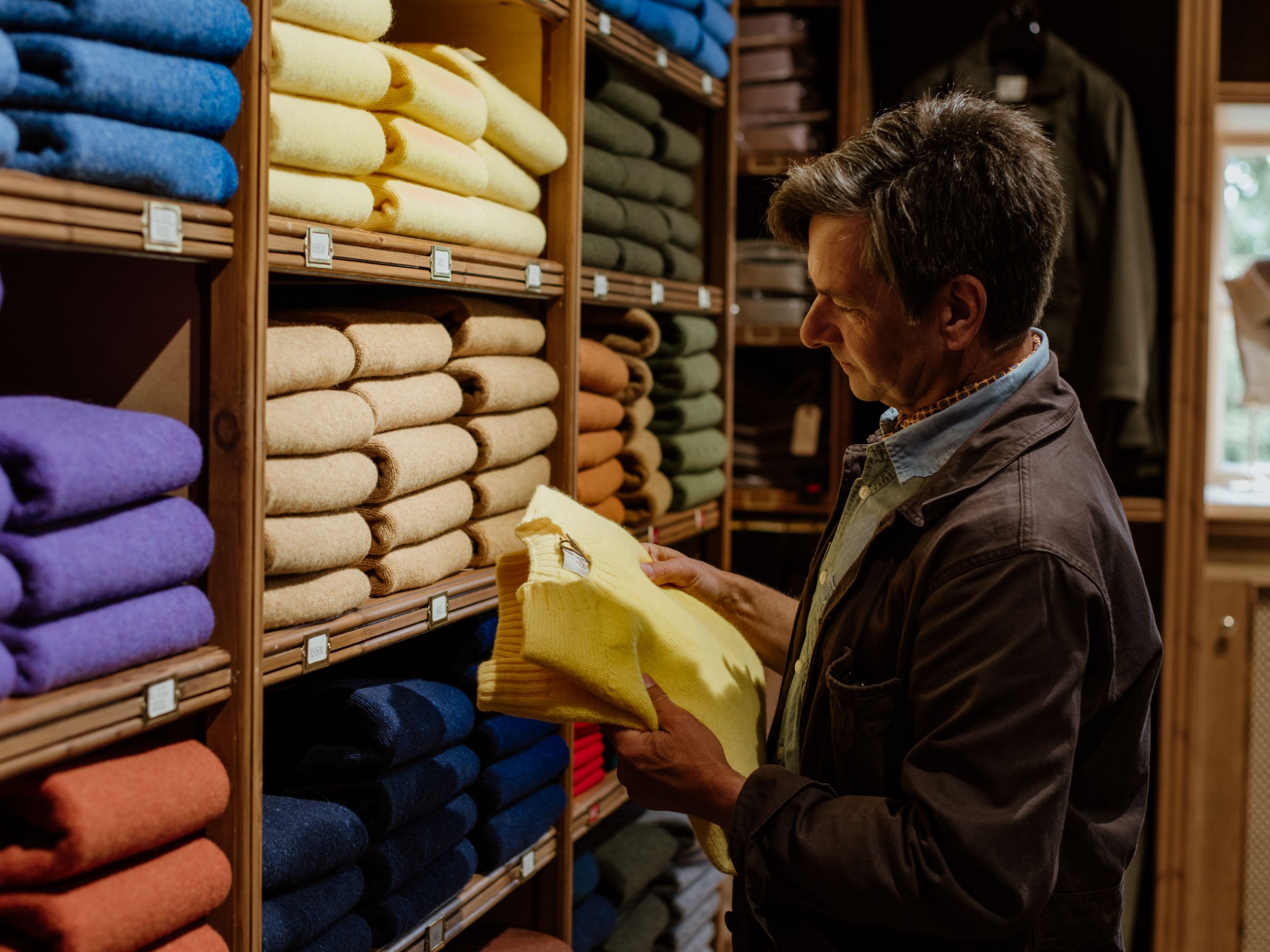 A person stands in front of neatly stacked shelves of folded sweaters in various colors, examining a yellow sweater in their hands inside a clothing store.
