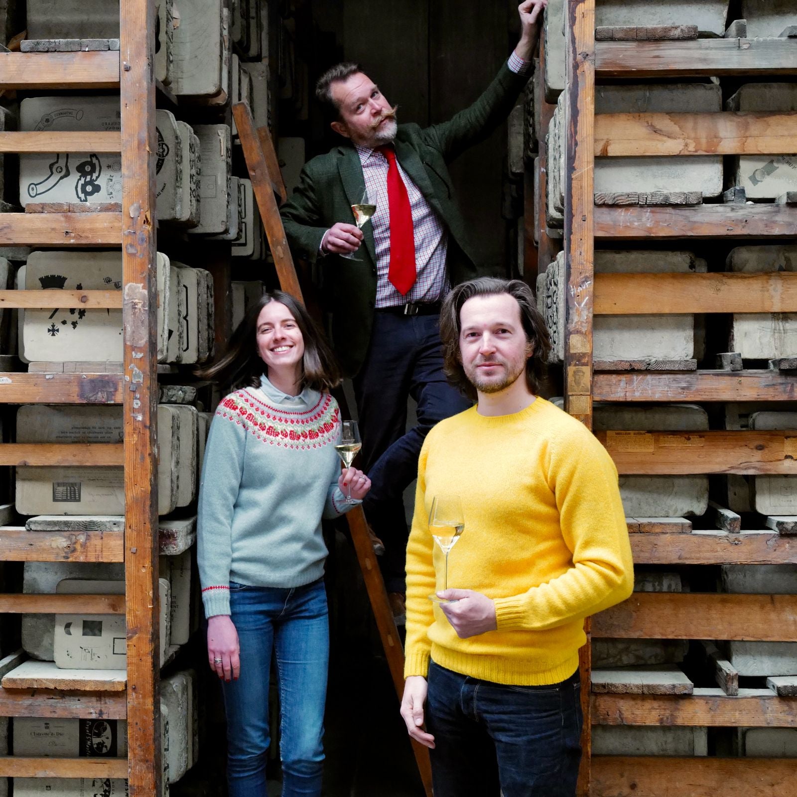 Three people holding glasses of white wine stand among tall wooden shelves filled with stacked vintage molds. One person stands on a ladder, while the other two pose in front, smiling at the camera.