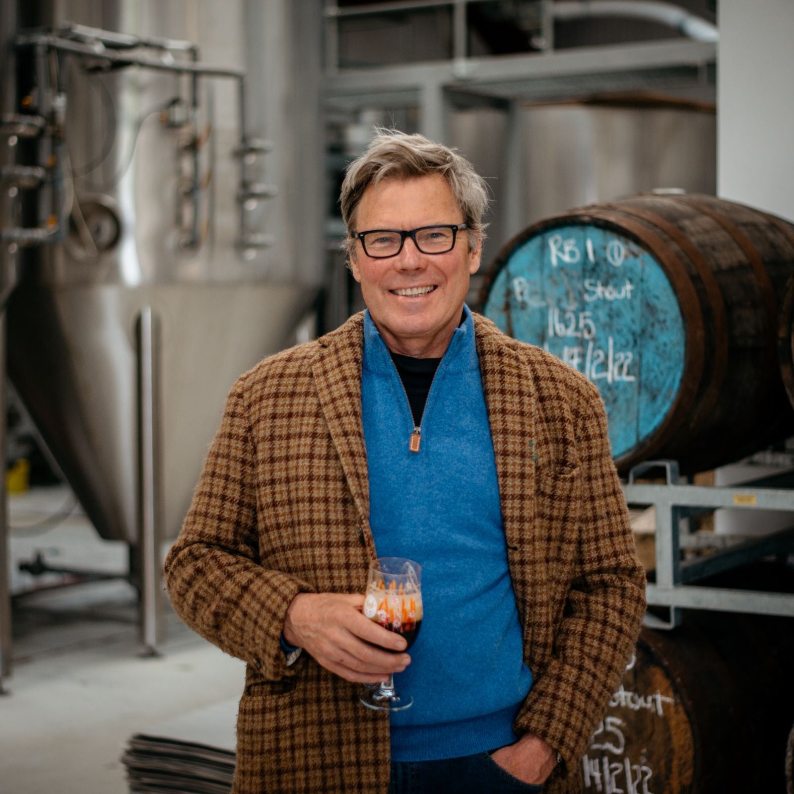 A smiling man in glasses and a checked brown jacket holds a glass of dark beer in a brewery, with large metal brewing tanks and wooden barrels in the background.