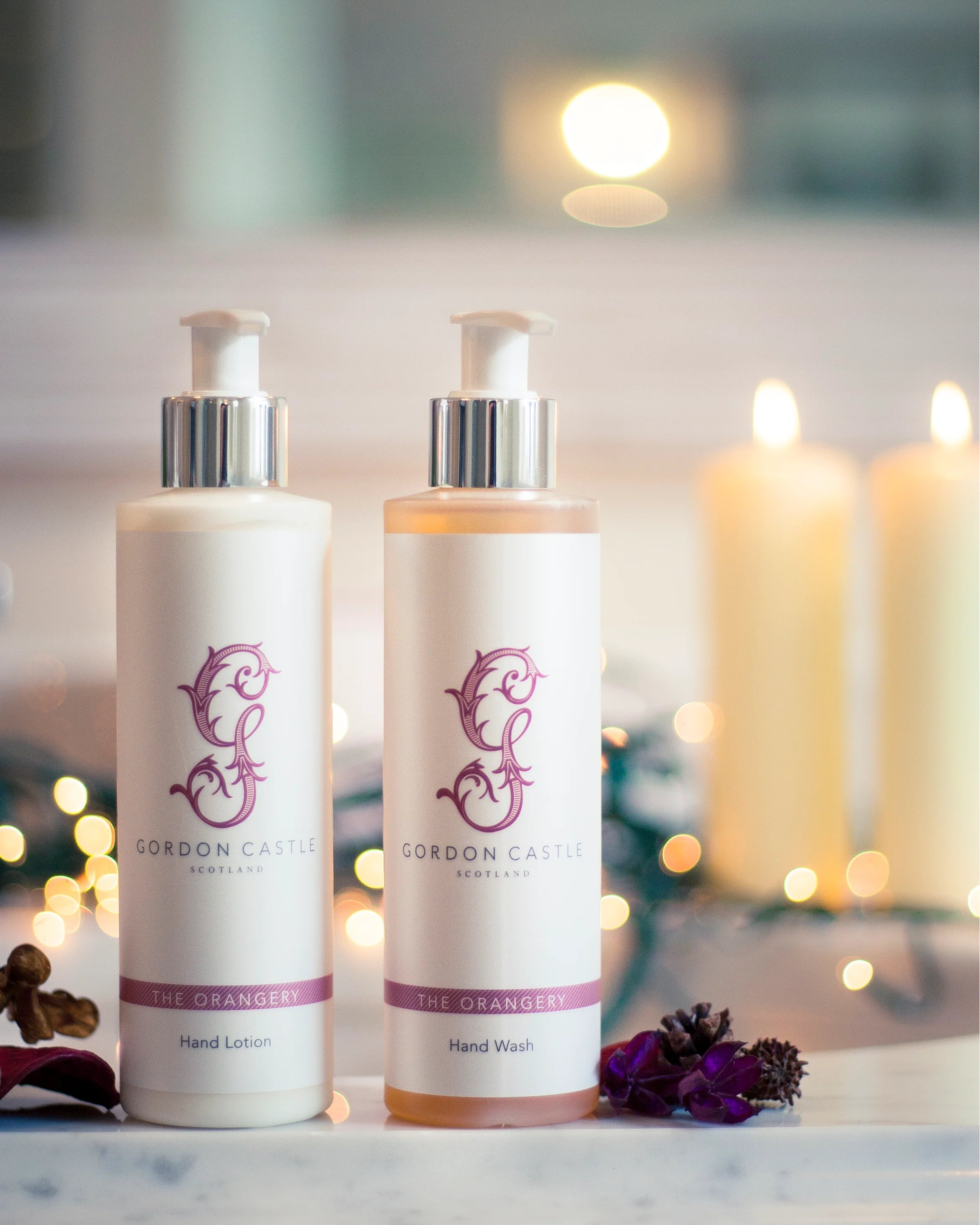 Two pump bottles labeled Gordon Castle Orangery Hand Lotion and citrus-scented Gordon Castle Hand Wash sit on a marble surface, with blurred candles and fairy lights in the background, creating a warm, cozy atmosphere.
