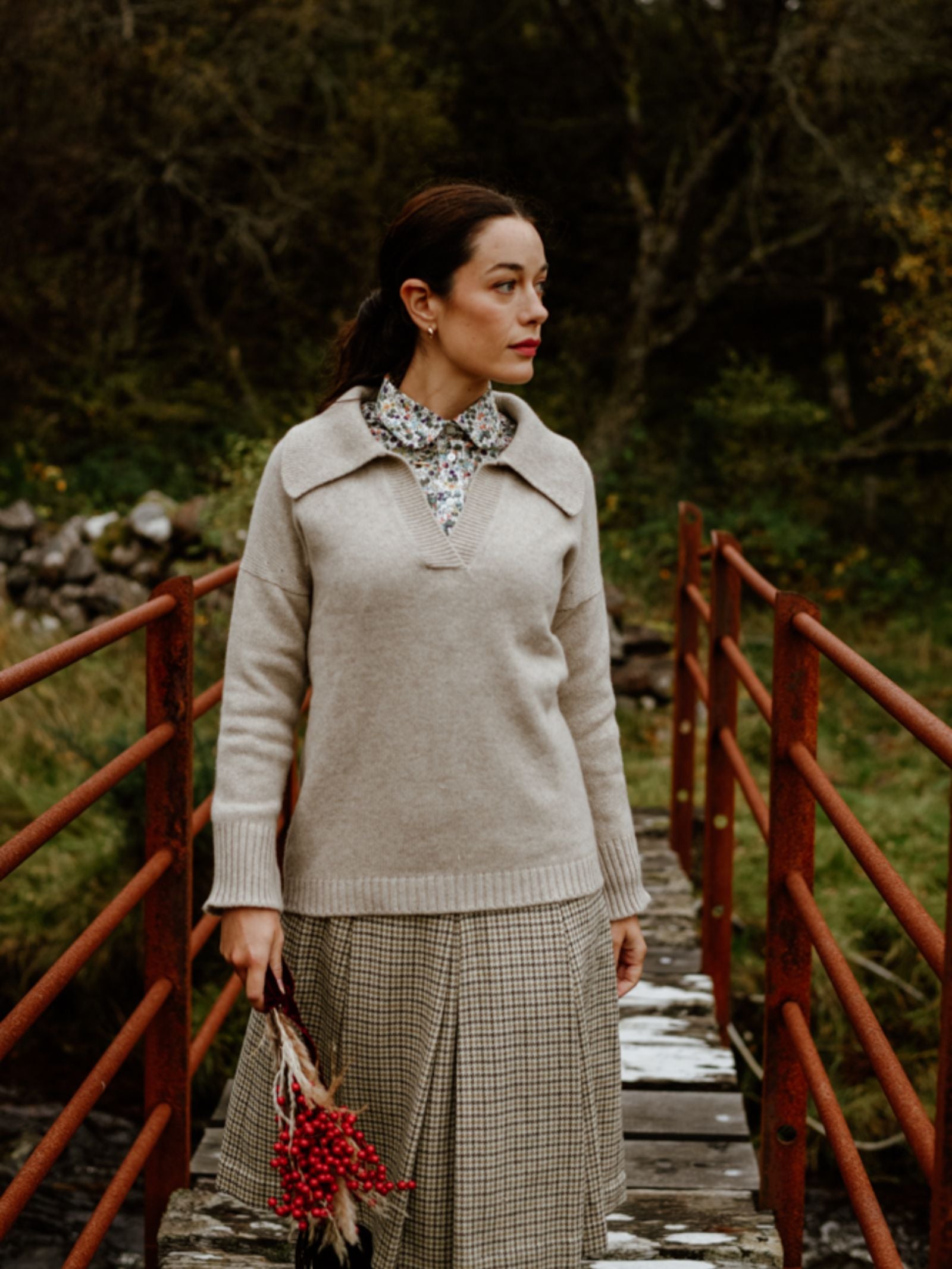 A woman in the Auld Stock Cashmere Shirt Jumper by Campbell's of Beauly, over a floral blouse and plaid skirt, stands on a red metal bridge holding red berries, with the lush Scottish Borders greenery behind her.