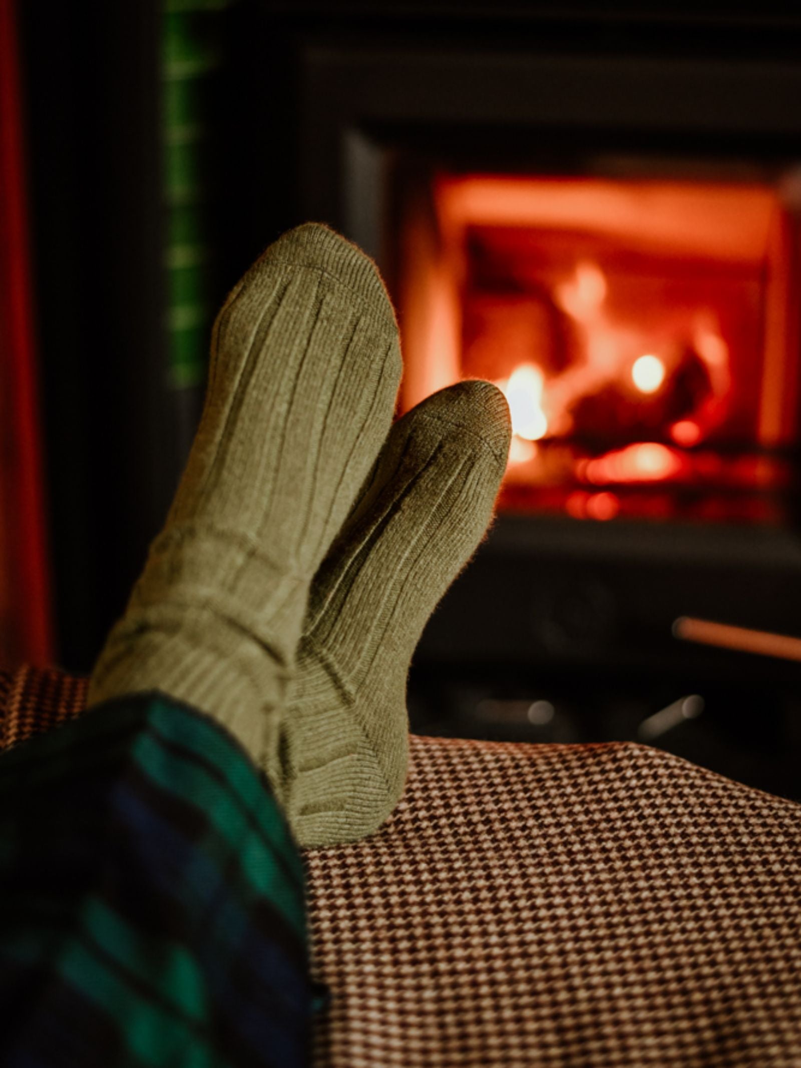 Wearing Campbell's of Beauly Rib Cashmere Socks, someone lounges in plaid pants by a glowing fireplace, evoking warmth and indoor comfort.