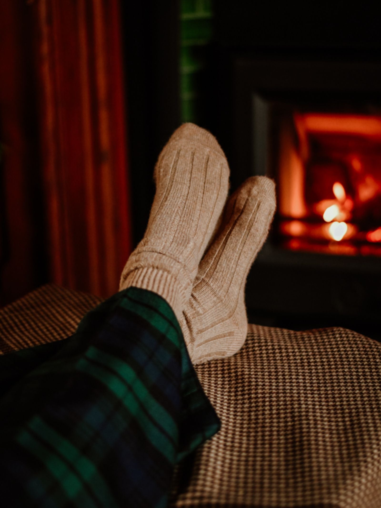 Wearing Campbell's of Beauly Rib Cashmere Socks and green plaid pajama pants, a person relaxes by the fireplace with feet propped on a checked blanket, soaking in the warmth of the fire.