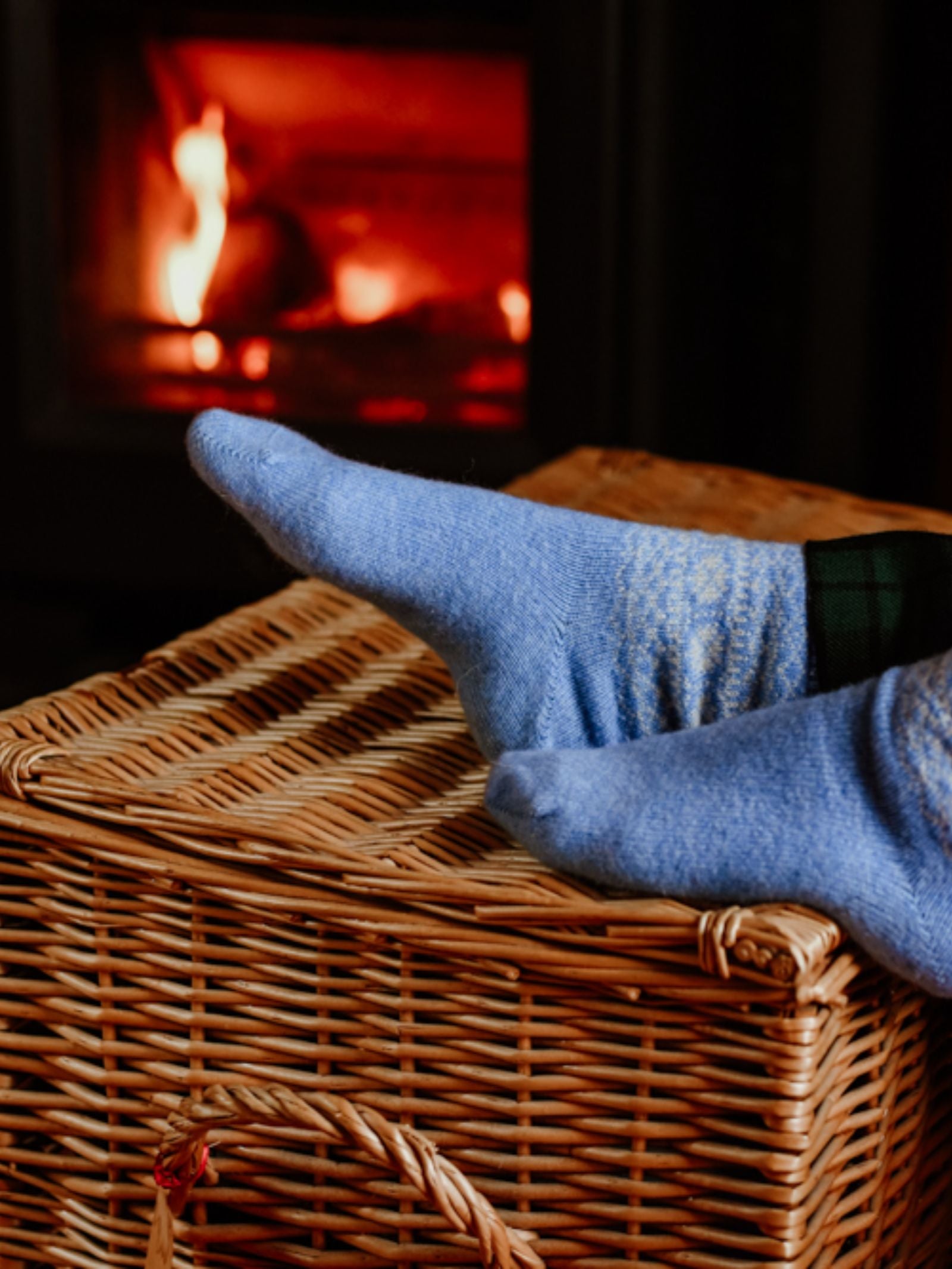 Someone wearing Campbell's of Beauly Two-Colour Fairisle Cashmere Socks rests their feet on a wicker basket by a glowing fireplace, creating a cozy and inviting atmosphere.