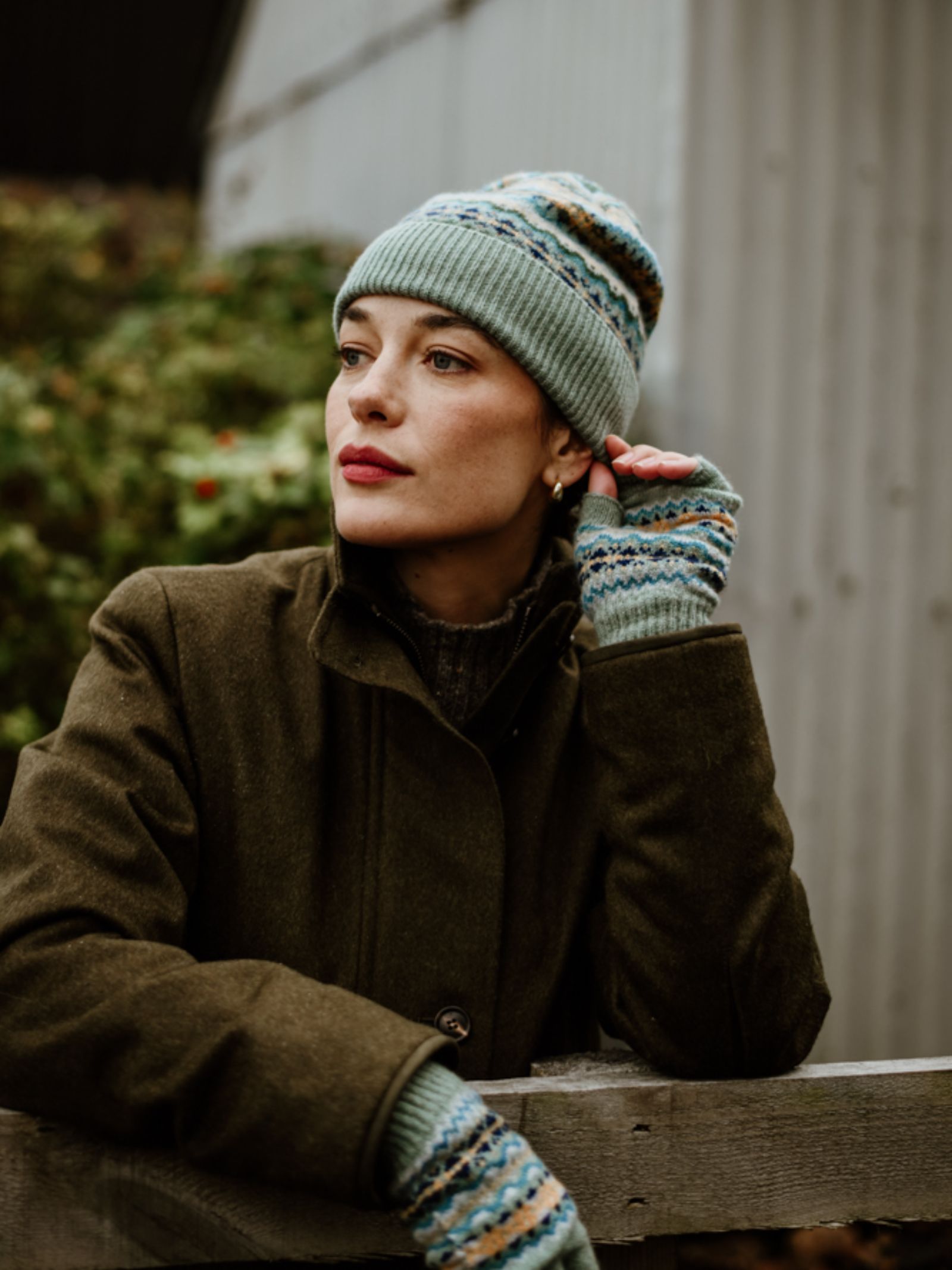 A person in a Campbell's of Beauly Auld Stock Lambswool Fairisle Hat leans on a wooden fence outdoors, dressed warmly among greenery with a metal building in the background—showcasing winter style.