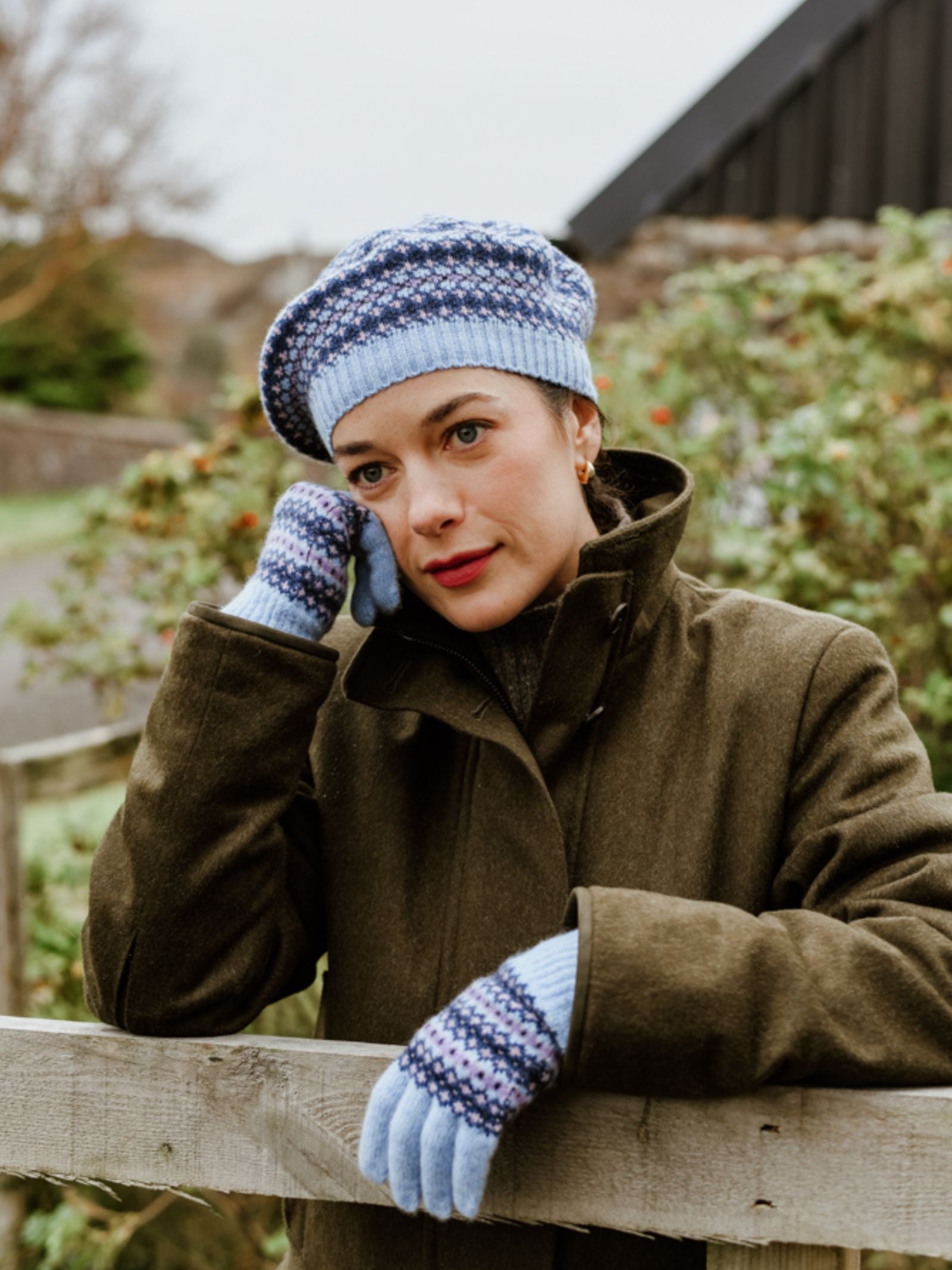 A woman in a green coat wears the Campbell’s of Beauly Fairisle Beret, along with matching gloves, as she leans on a wooden fence outdoors, resting her head on her gloved hand and gazing thoughtfully into the distance.