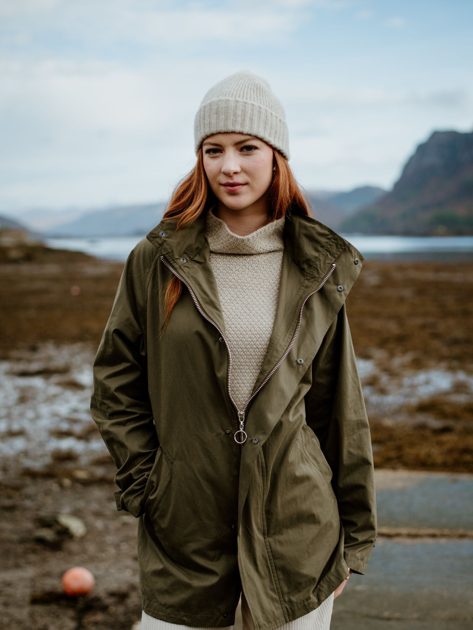 A woman with long red hair wears the Campbell's of Beauly Cream Lambswool Hat, a light sweater, and an olive green jacket while standing by a rocky shoreline in the Scottish Borders with mountains and water behind her.