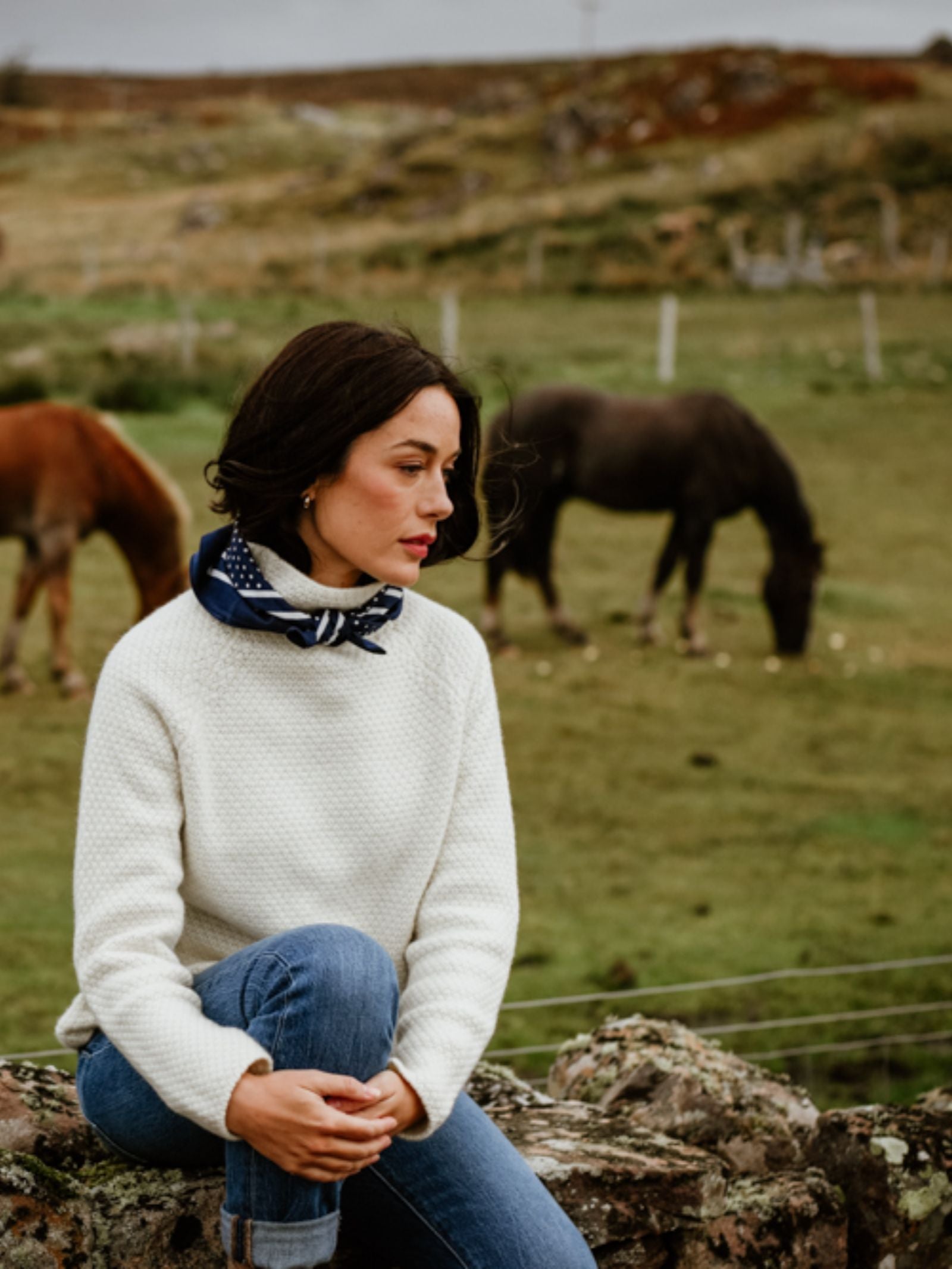 A woman with dark hair wears the Campbell's of Beauly Cashmere Funnel Neck Jumper in white, paired with blue jeans and a blue scarf, as she sits on a stone wall in a grassy field with two horses grazing peacefully in the background.