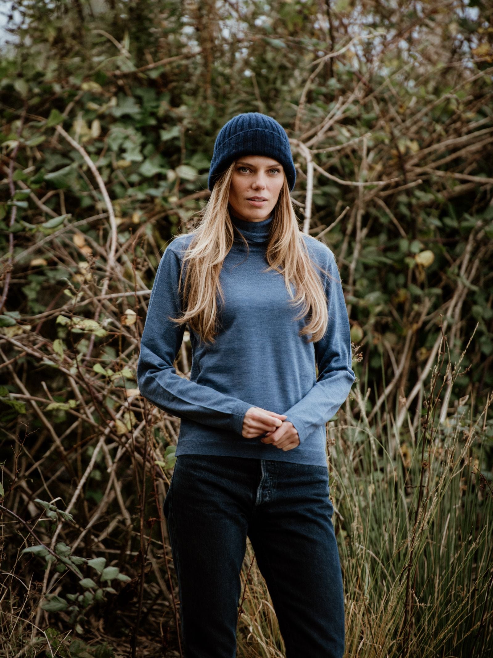 A woman with long blonde hair in a blue beanie and the Auld Stock Merino Poloneck by Campbell's of Beauly stands outdoors before tall, dense greenery and branches.
