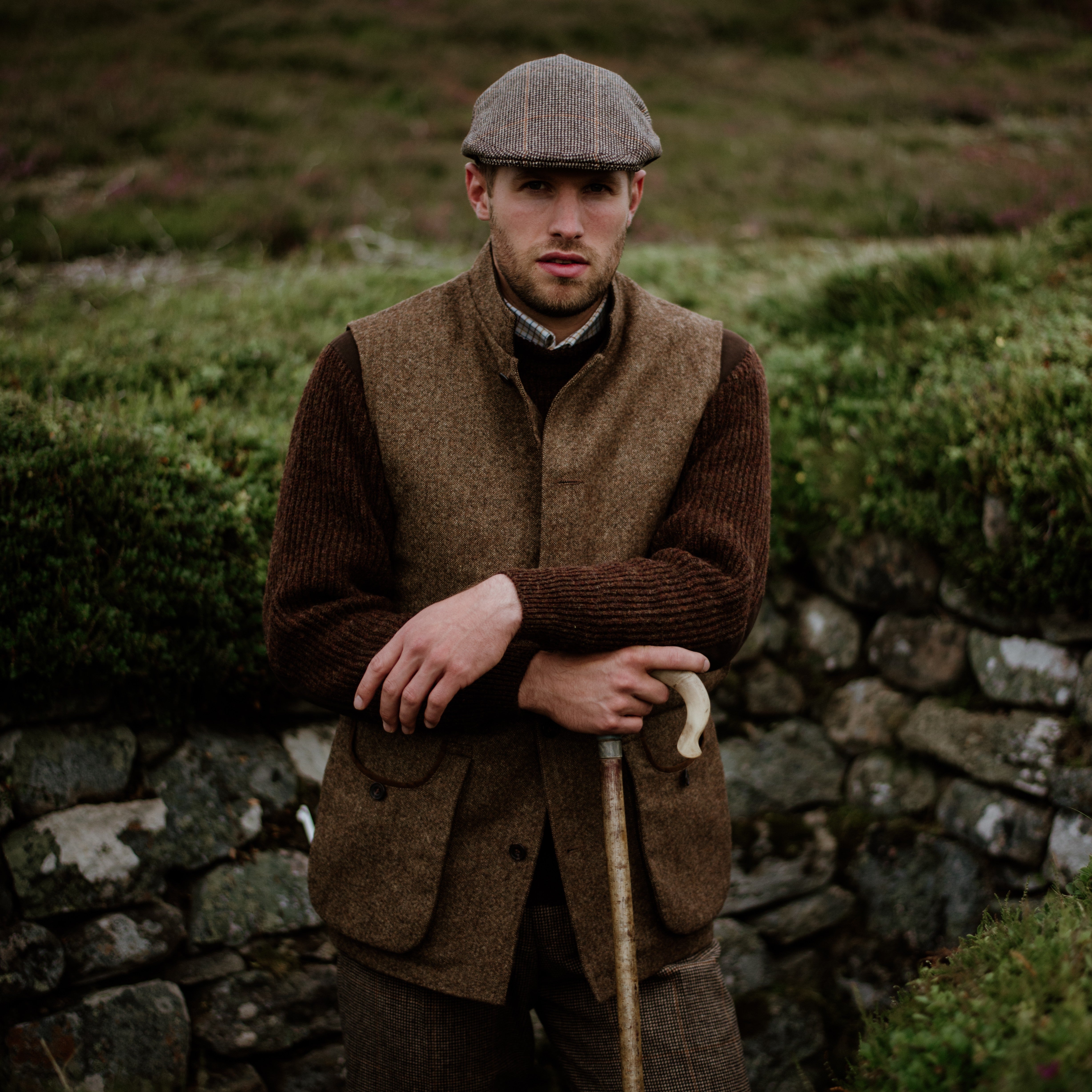 A man in vintage country attire, including a flat cap, brown vest, and sweater, stands outdoors by a stone wall, holding a walking stick with greenery in the background.