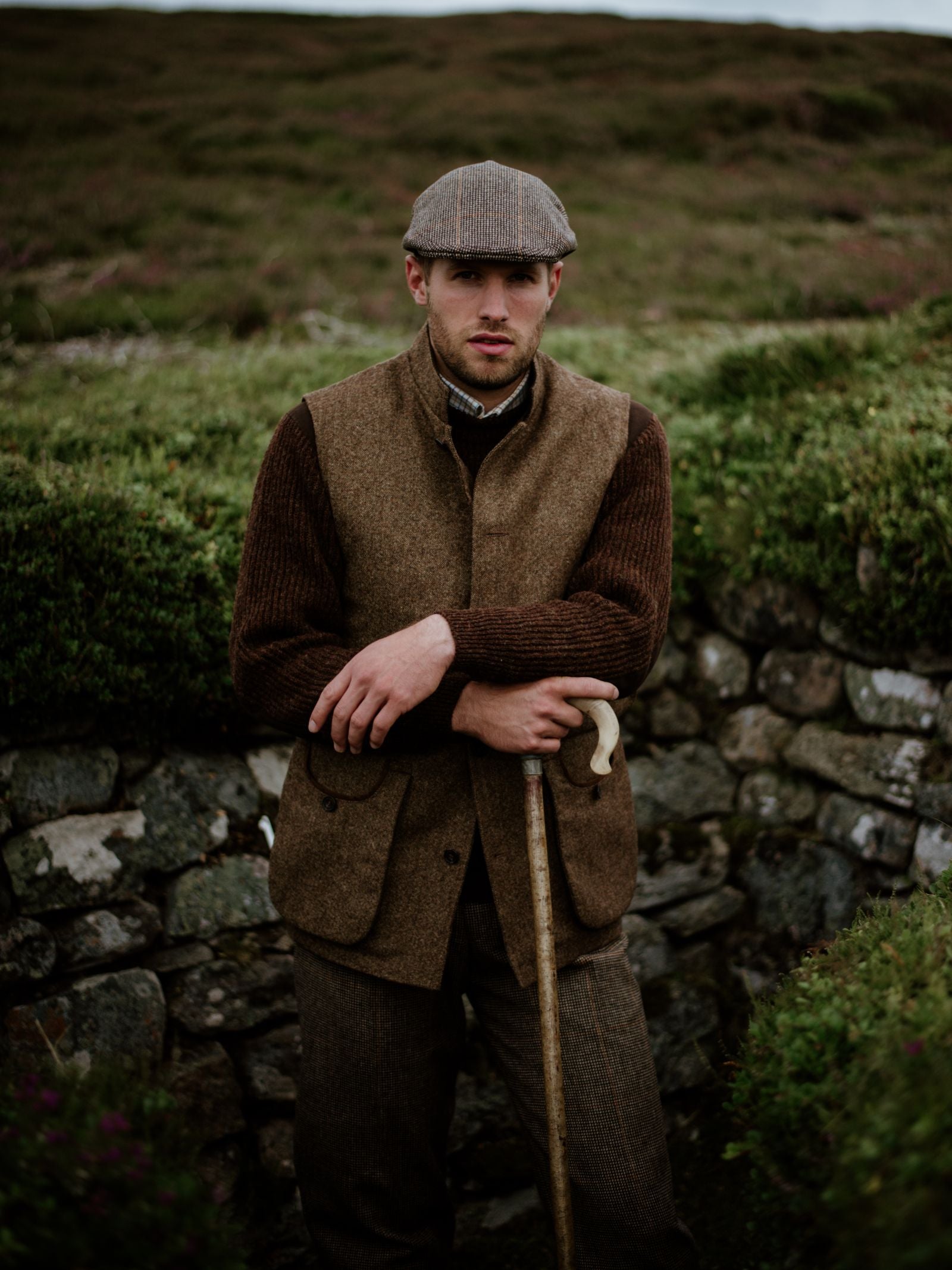 A man wearing the Campbell's of Beauly Auld Stock Field Nehru Gilet and a flat cap stands by a stone wall outdoors, holding a walking stick, with green shrubs and grass in the background.