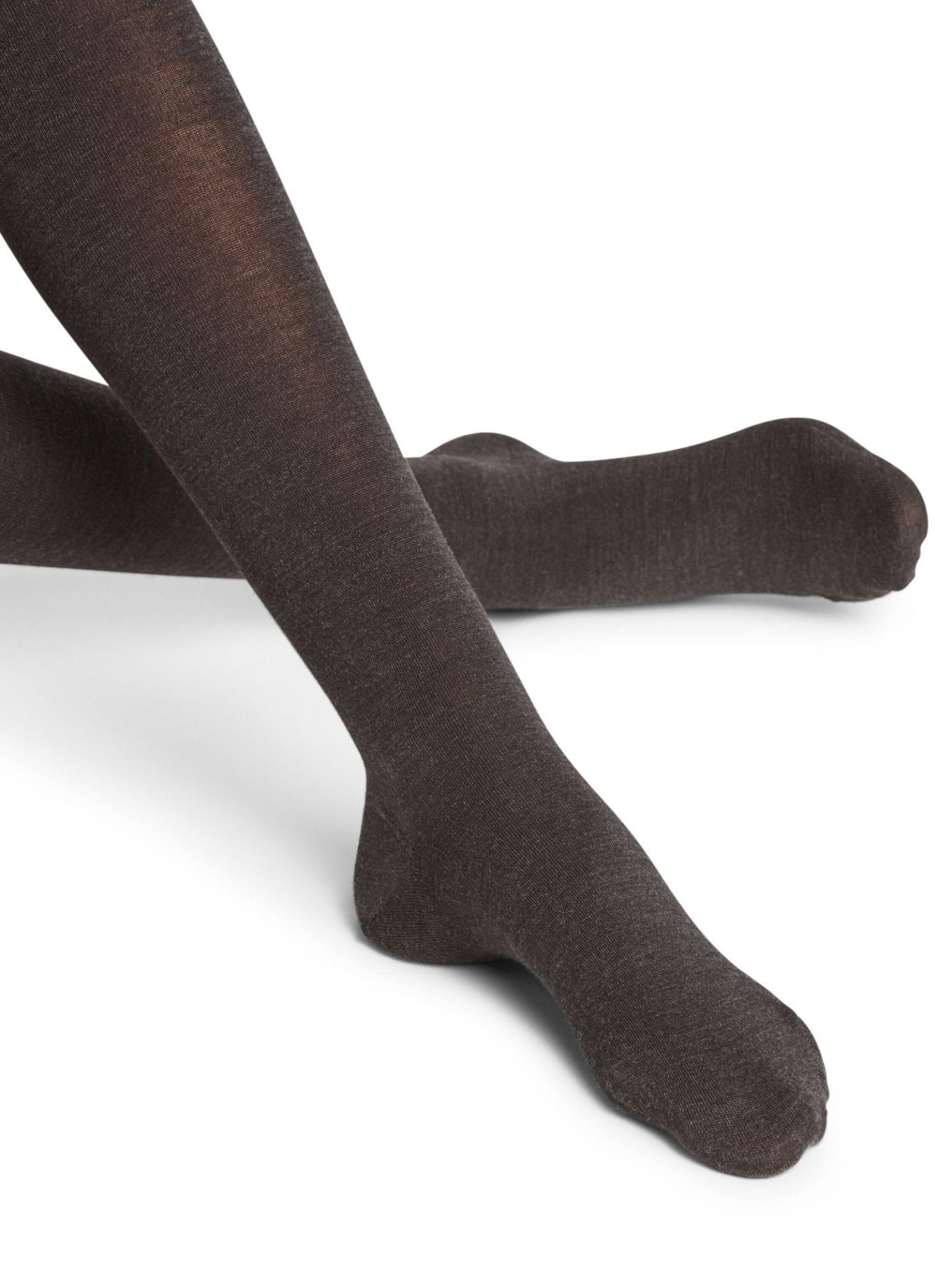 A person wearing FALKE Merino Tights in dark brown sits cross-legged on a white background, highlighting the lower legs and feet.