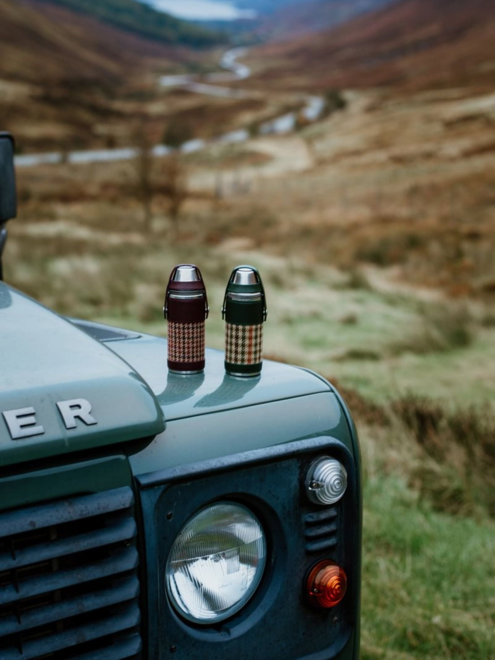Two patterned thermoses, including the 8oz Bordeaux Hunter Flask from Campbell's of Beauly x Ettinger, rest on the hood of a green Land Rover in a grassy, hilly landscape.