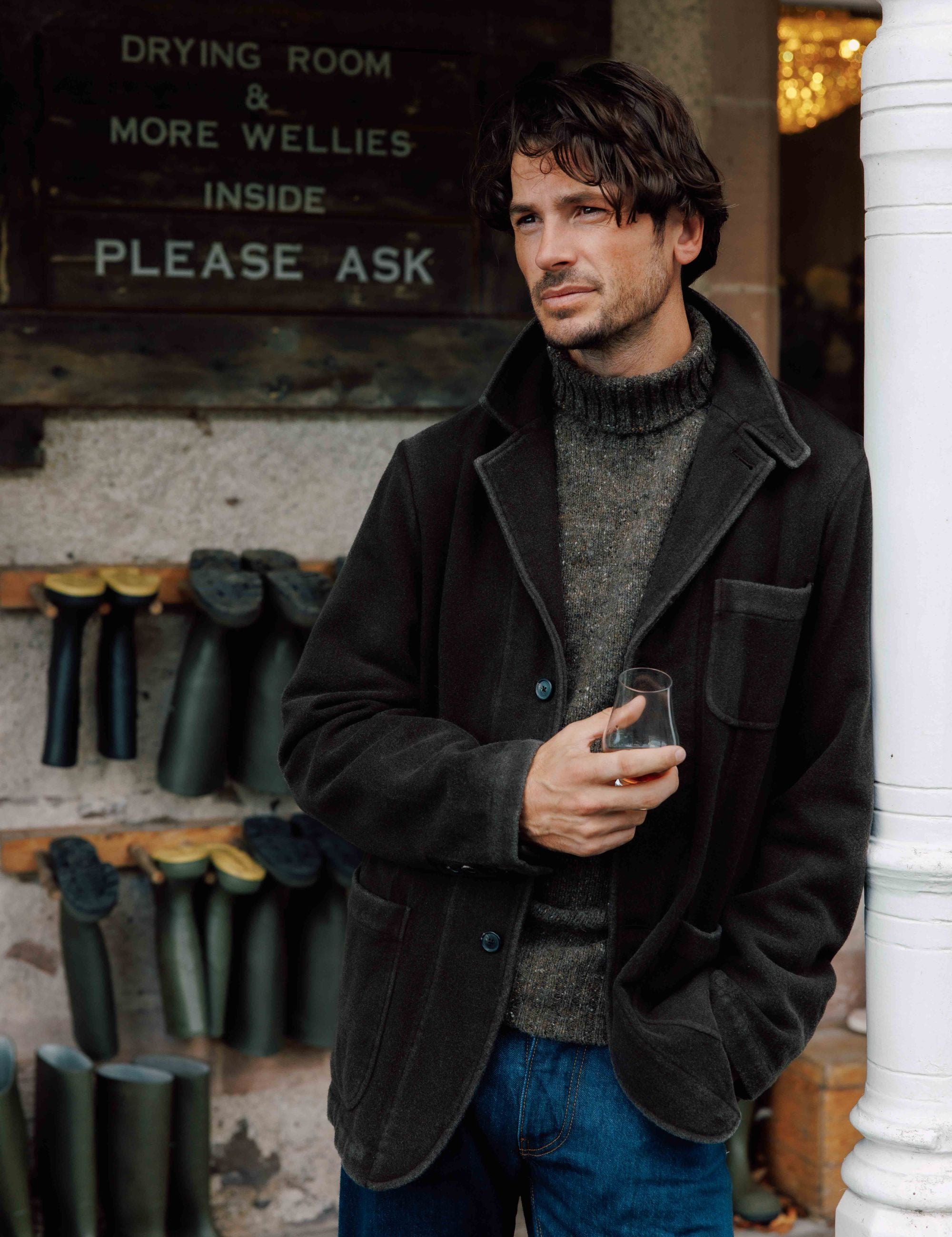 A man in a dark coat holds a drink and stands by a white post, with racks of rubber boots on the wall behind him and a sign reading “DRYING ROOM & MORE WELLIES INSIDE PLEASE ASK.”.