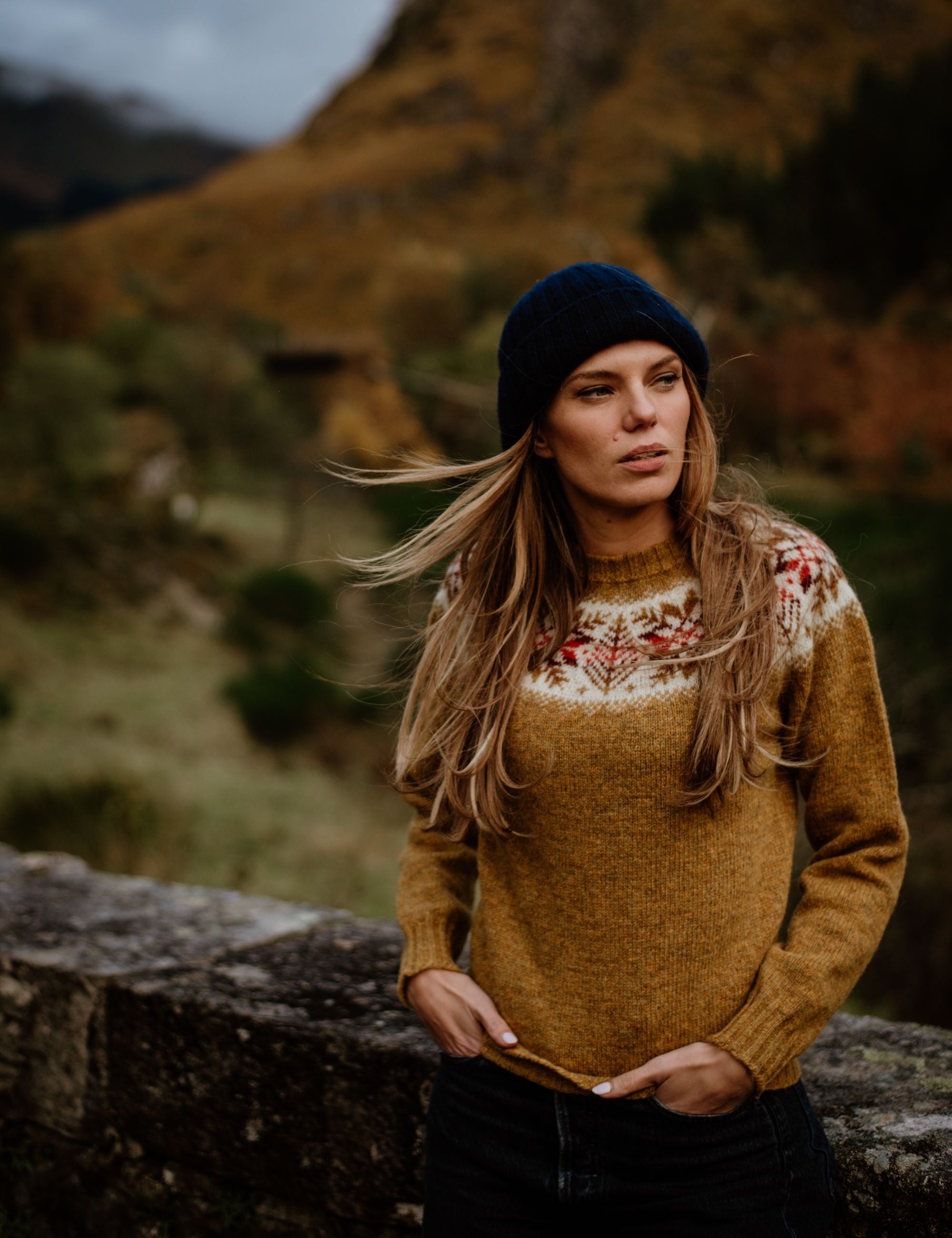 A woman with long blonde hair, wearing a mustard yellow sweater with a patterned yoke and a navy knit hat, stands outdoors by a stone wall, with hills and trees in the blurred background.