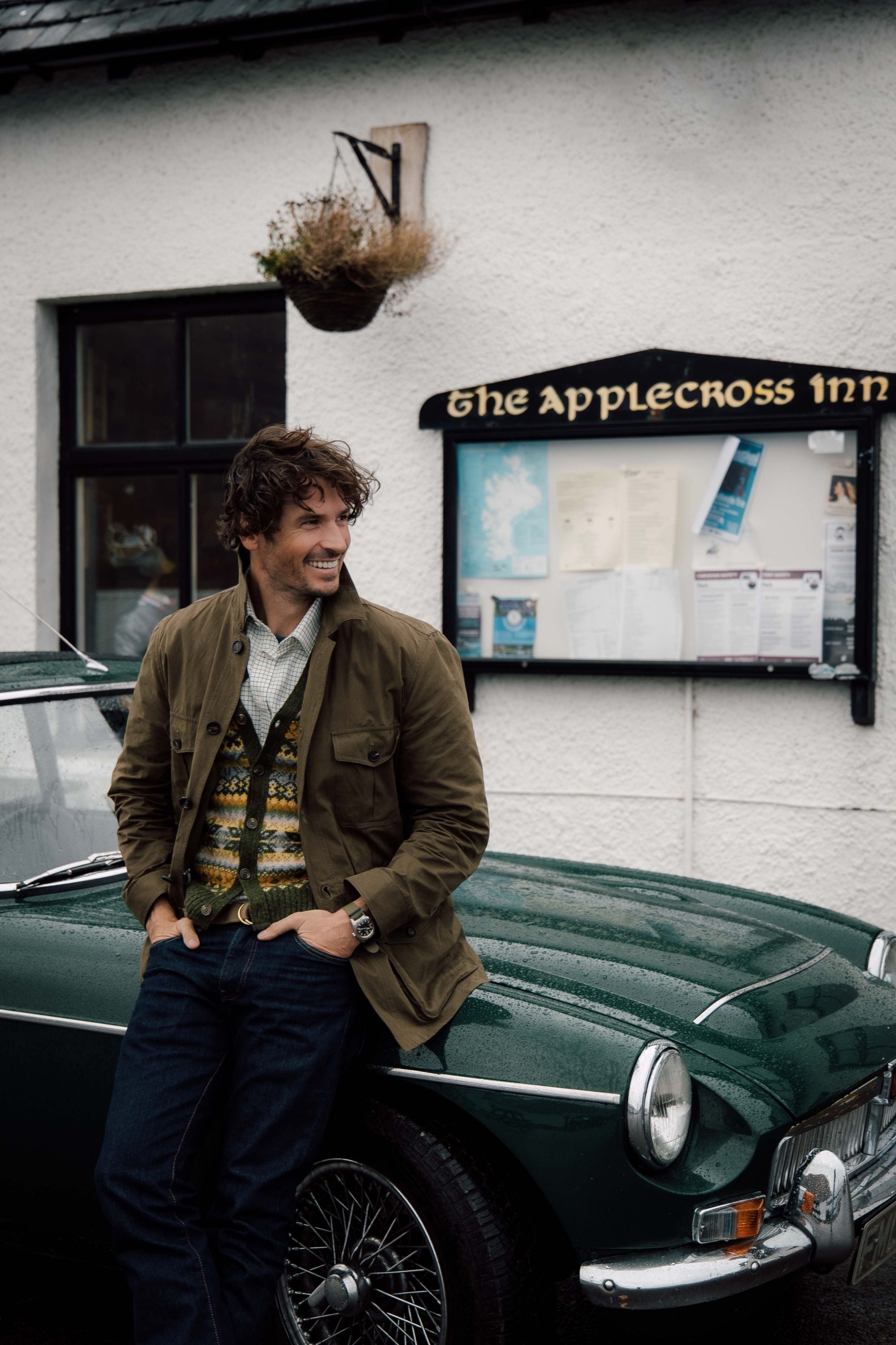 A man in a brown jacket and patterned sweater smiles while leaning against a classic green car parked outside The Applecross Inn, with a map and notices displayed on the wall behind him.
