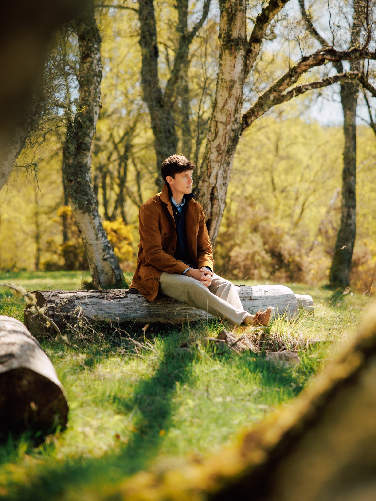 A man in a brown jacket and beige pants sits on a log in a sunlit forest clearing, surrounded by trees and green grass, looking thoughtful.