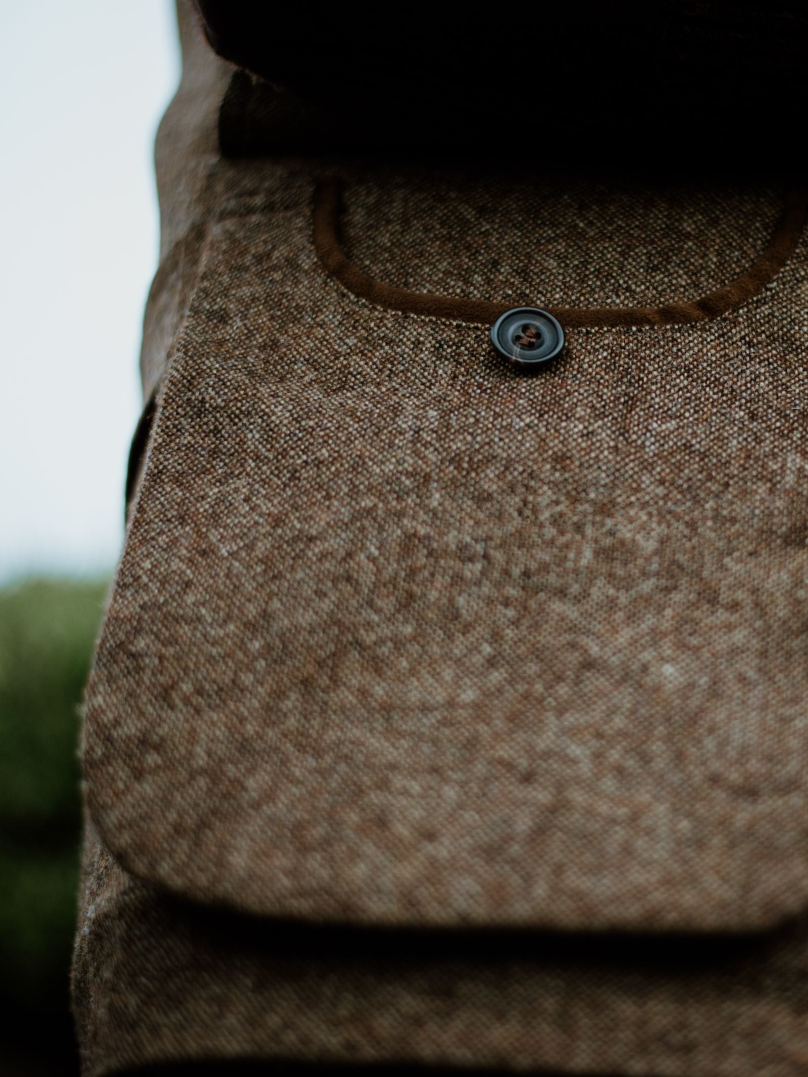 Close-up of the Auld Stock Field Nehru Gilet by Campbell's of Beauly, highlighting its textured brown tweed, detailed stitching, and black button. Inspired by British moleskin, the expert craftsmanship stands out against a softly blurred green backdrop.