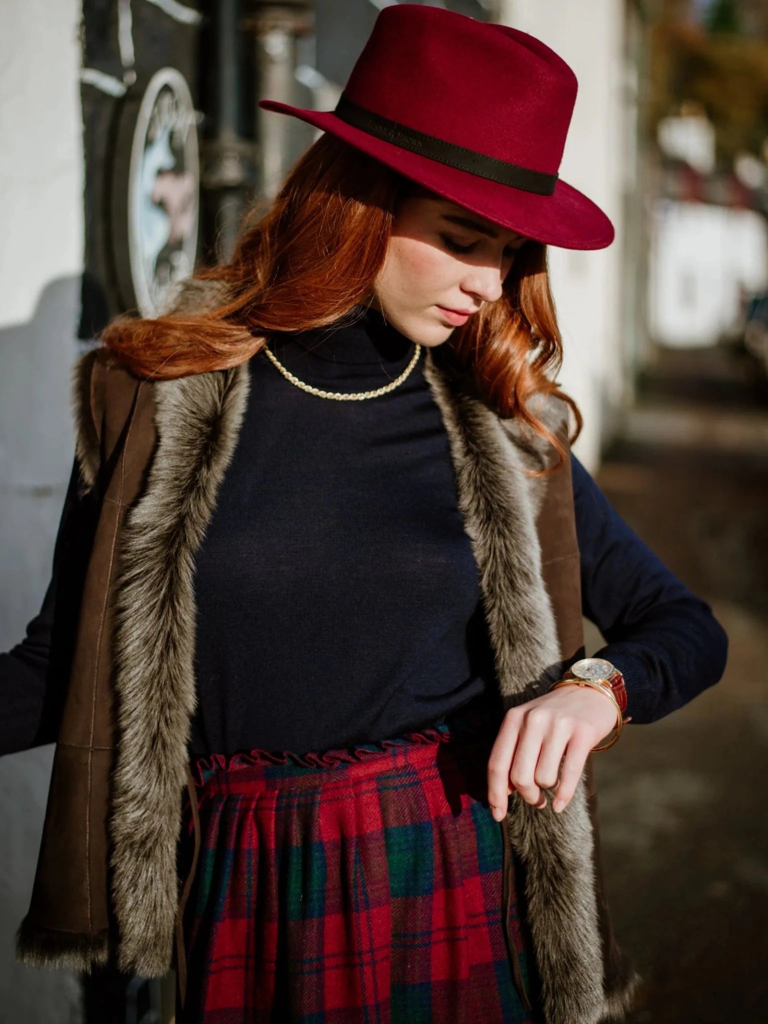 A woman with long red hair wears a Hicks & Brown Auld Stock Gamebird Fedora with a feather wrap, paired with a black turtleneck, faux fur vest, and plaid skirt. She checks her wristwatch while standing outdoors in natural light.