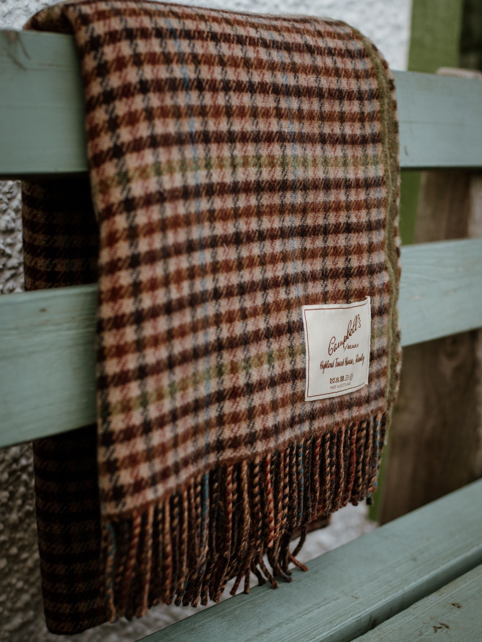 A Double-Faced Lambswool Throw by Campbells of Beauly, featuring brown, green, and red classic checks with fringed edges, is draped over a light blue wooden bench. The blanket has a “Campbells of Beauly” label attached.