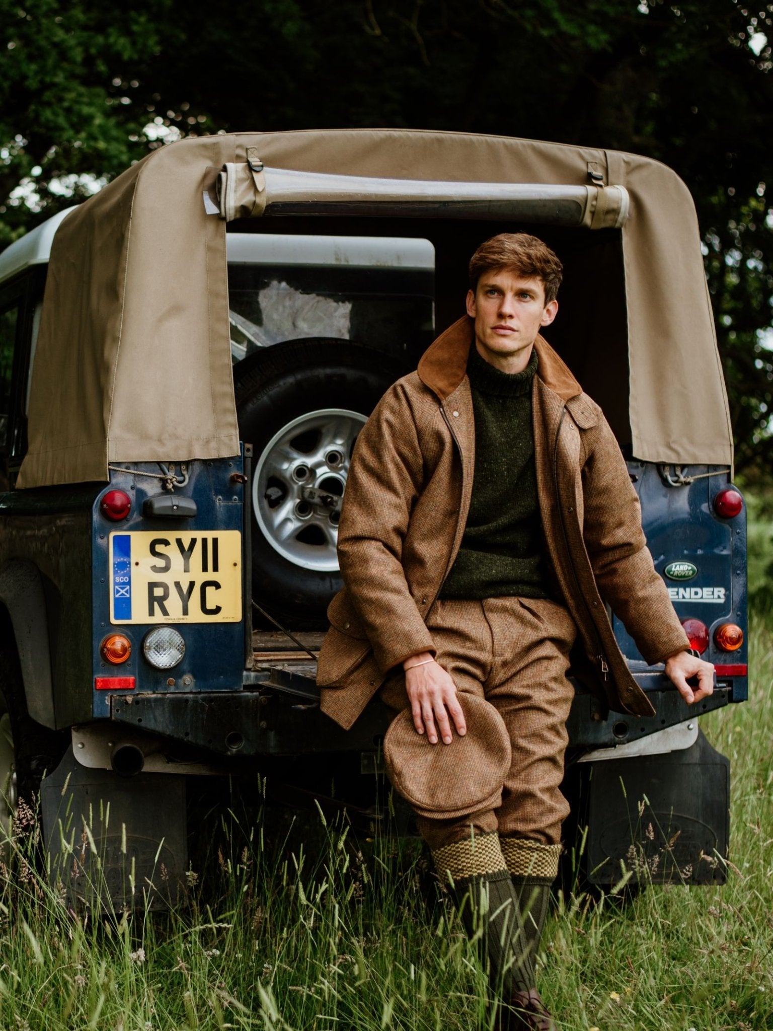 A man in a Campbell's of Beauly Tweed Field Coat sits on the open back of a UK-made Land Rover Defender with a beige canopy in a grassy field, holding his hat and gazing into the distance, with the license plate visible.