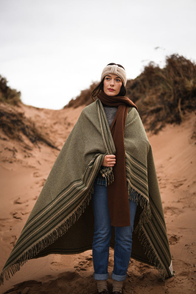 A woman stands on a sandy path between dunes, wrapped in a large checkered blanket. She wears jeans, a brown scarf, a gray sweater, and a knitted headband, looking ahead with a calm expression.