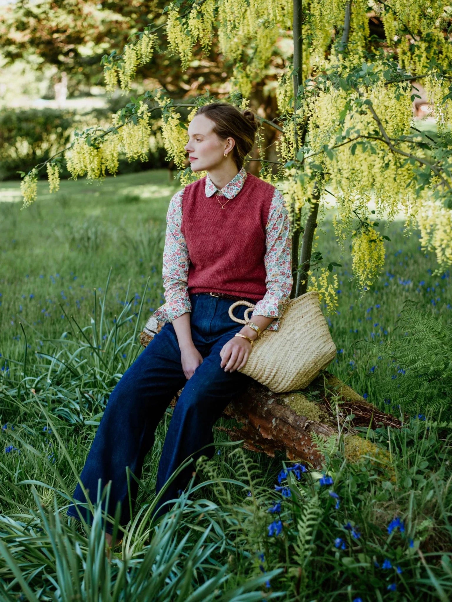 A woman sits on a log in a grassy garden, wearing Campbell's of Beauly Liberty Print Cotton Shirt beneath a red sweater vest and wide-leg jeans. She holds a woven straw bag, surrounded by greenery and yellow flowers.